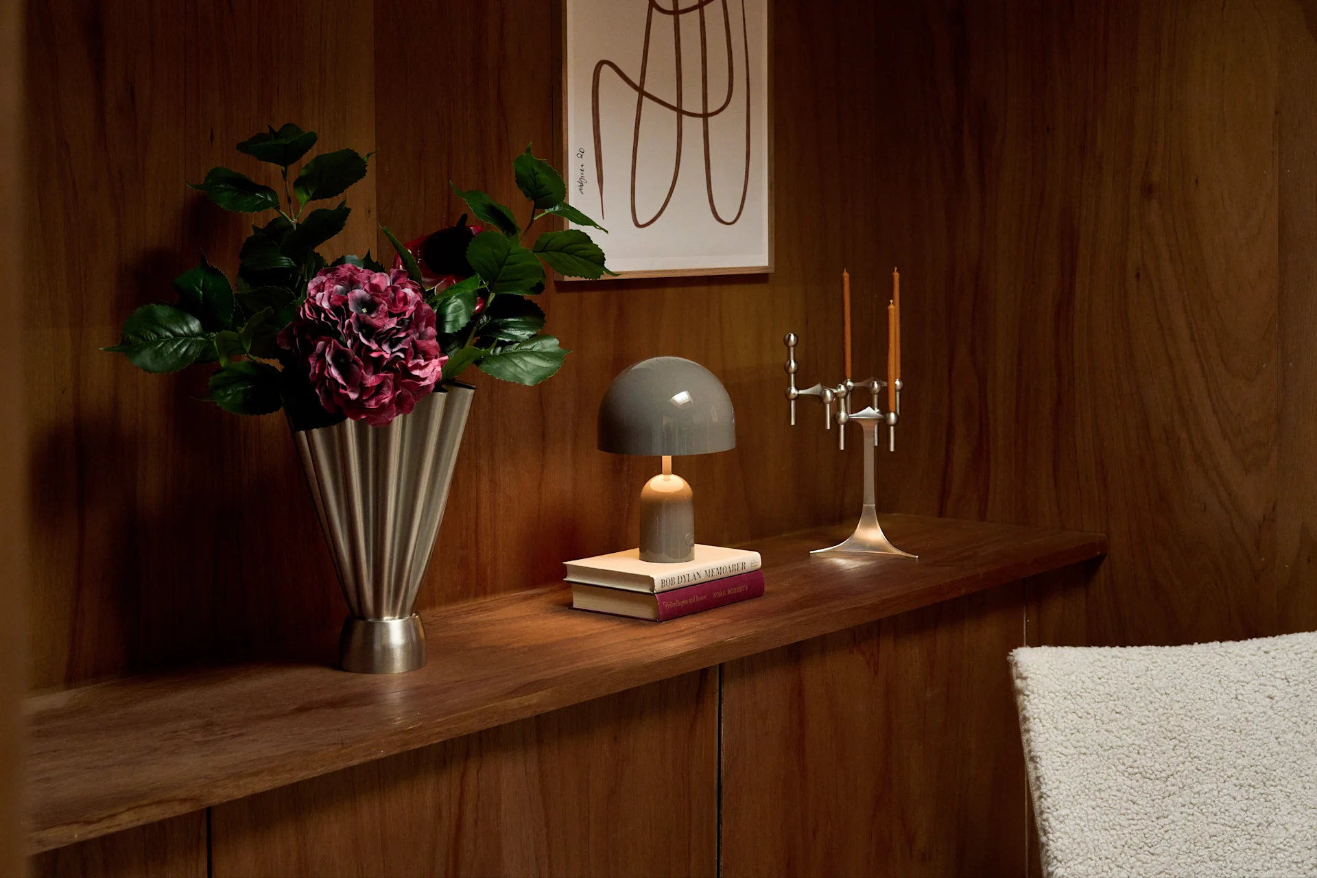 Estante de madera: jarrón estriado plateado con flores moradas, lámpara gris sobre libros, y candelabro plateado con dos velas.