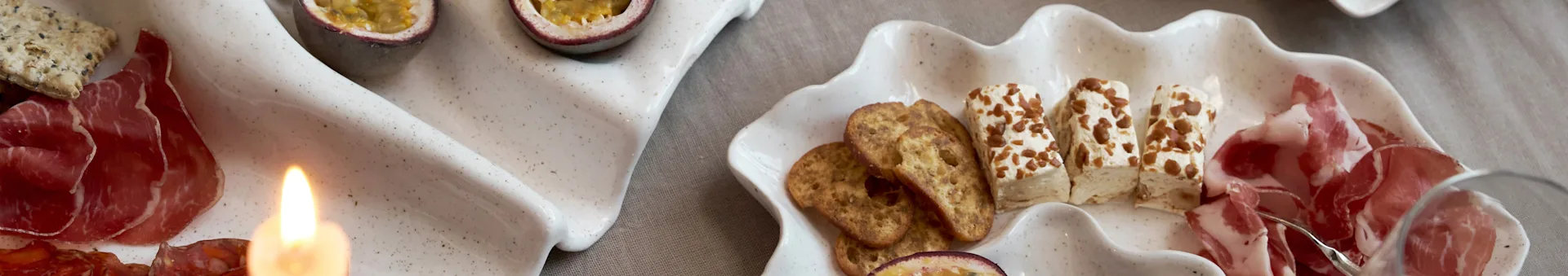 Tabla de aperitivos en dos bandejas en forma de concha con embutidos, galletas saladas, queso y maracuyá, con una vela encendida.