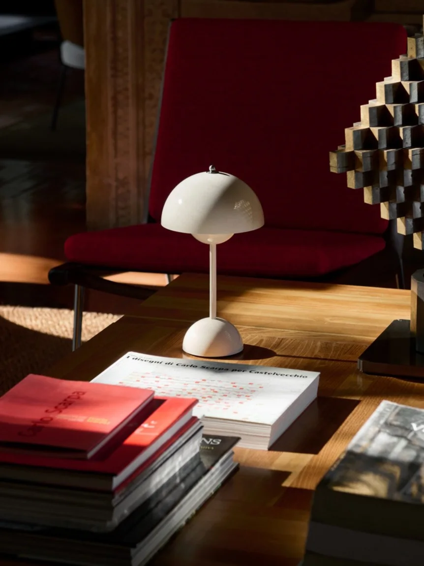 Una lámpara de mesa blanca con forma de hongo, libros y una escultura geométrica en una mesa de café de madera, con un sillón rojo detrás.