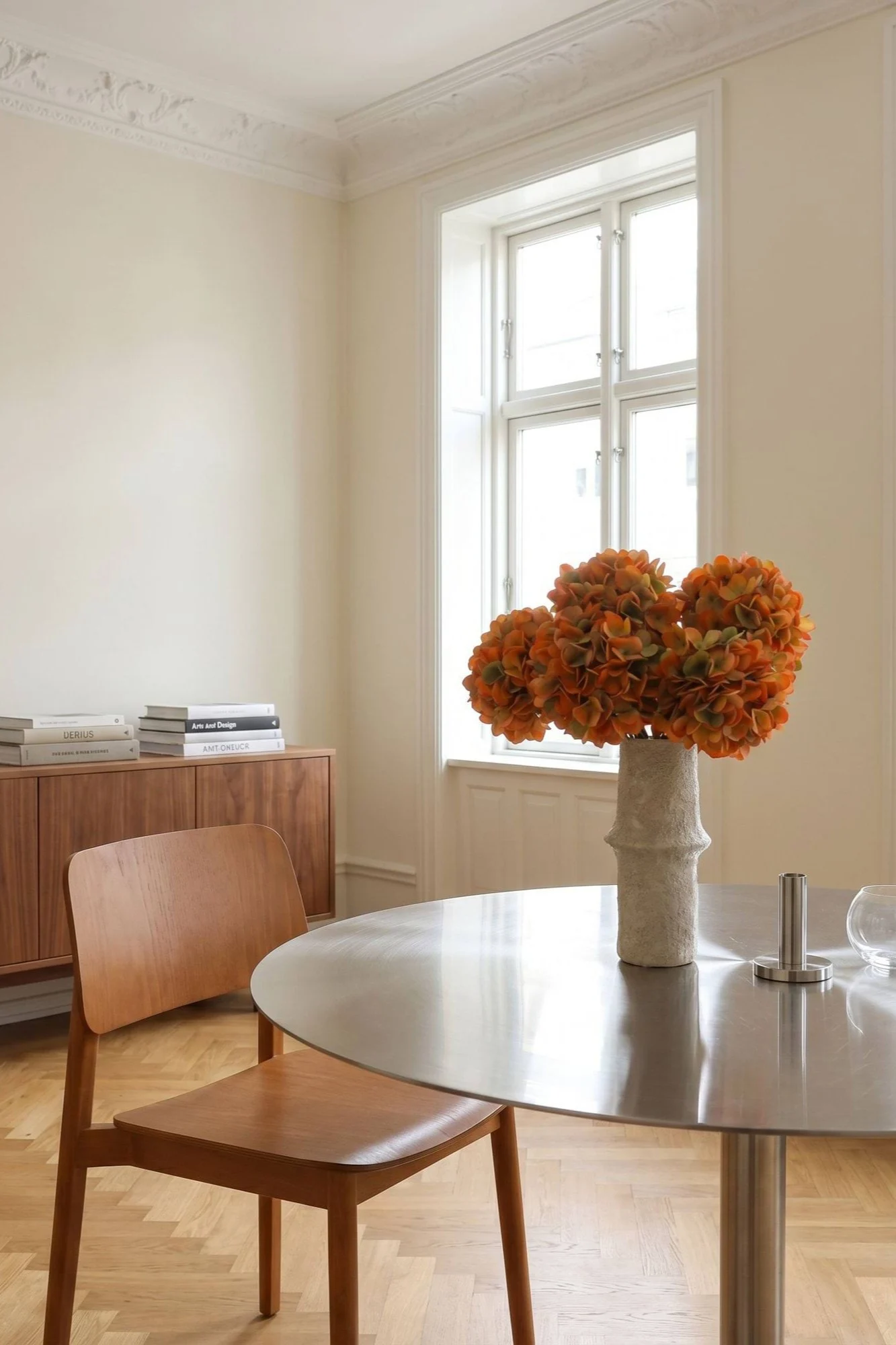 Un luminoso comedor con una mesa redonda de metal, una silla de madera y un jarrón de flores naranjas.