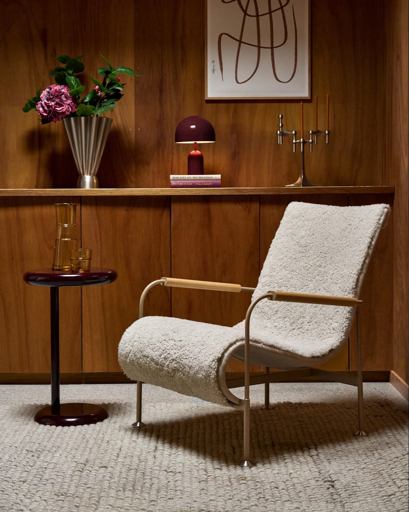 Habitación con paneles de madera, sillón mullido, mesa auxiliar roja, estante con jarrón, lámpara, candelabro y arte abstracto.