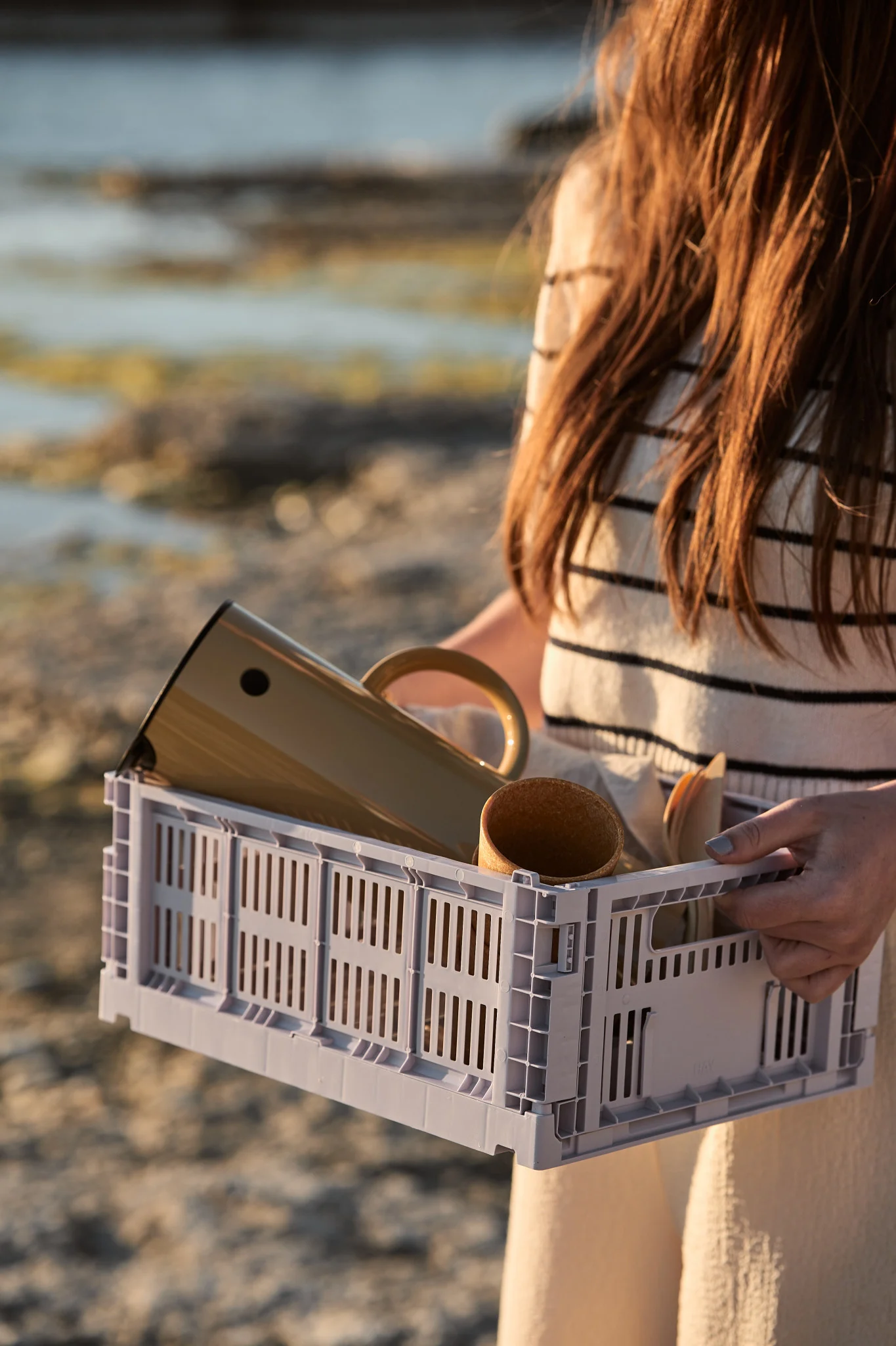 Una persona sostiene una caja de listones de color gris claro que contiene un termo de color beige, una taza y cubiertos de madera, con la playa al fondo.