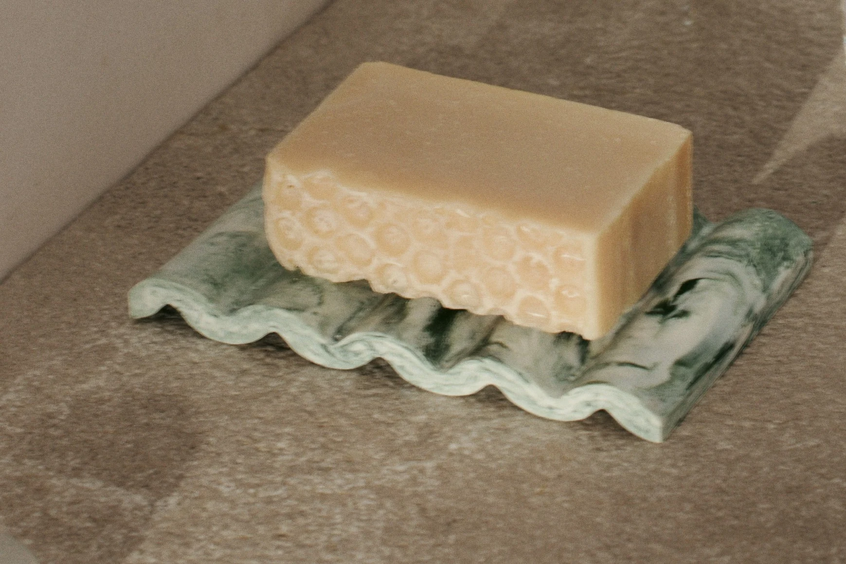 Pastilla de jabón beige claro con textura de panal en una jabonera ondulada de mármol verde.