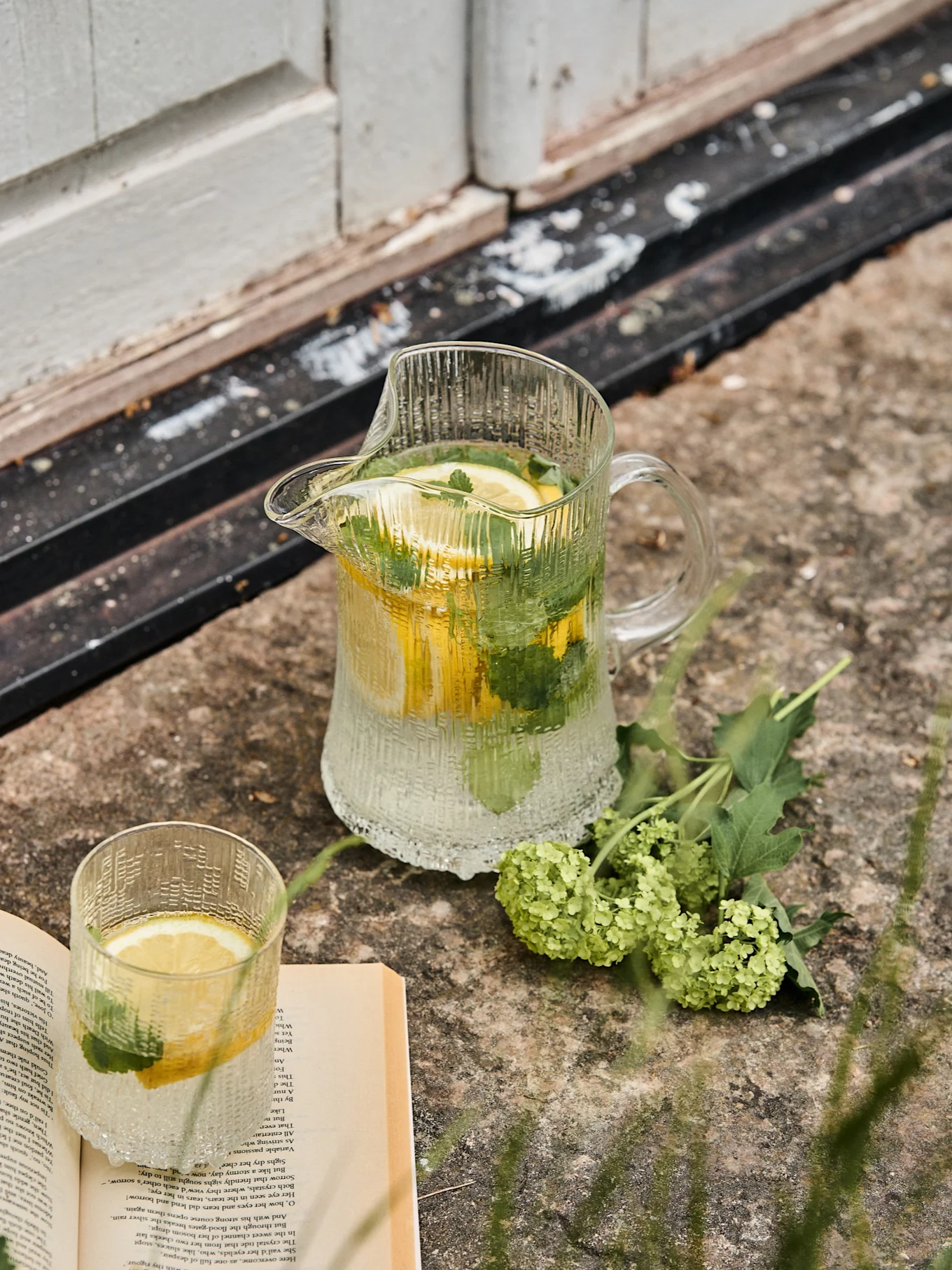 Jarra de vidrio acanalado y vaso a juego con agua de limón y menta, junto a un libro abierto y flores verdes sobre piedra.