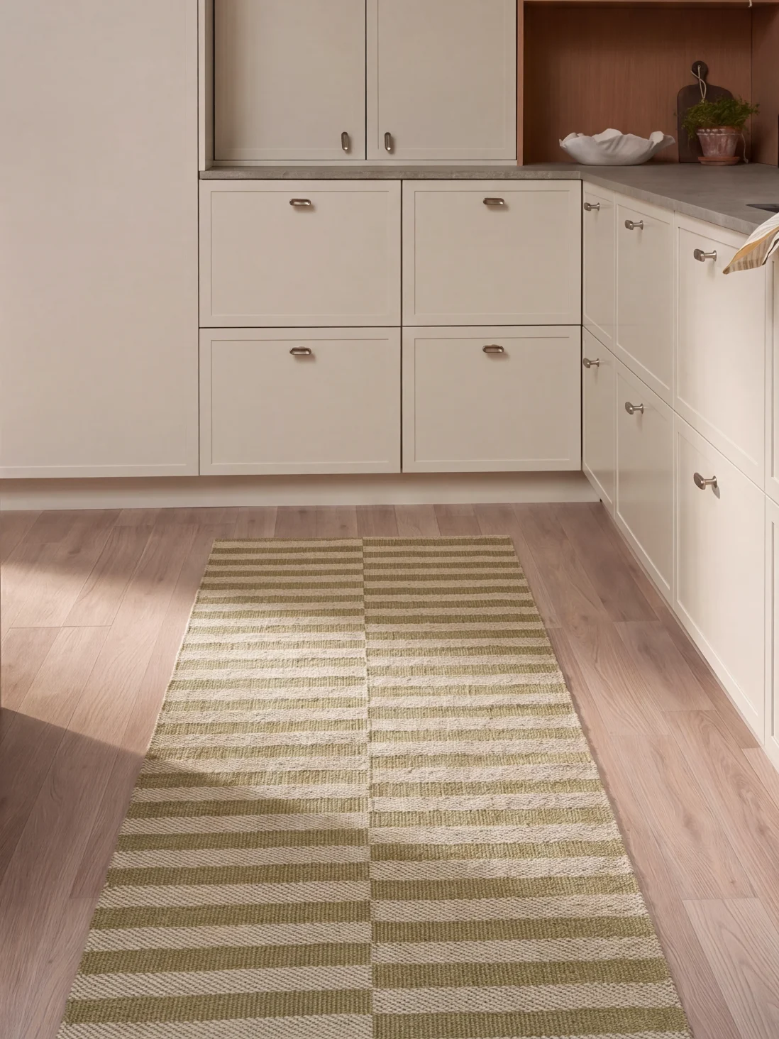 Una cocina con armarios claros, suelo de madera y una alfombra larga rectangular con rayas horizontales verdes y blancas.