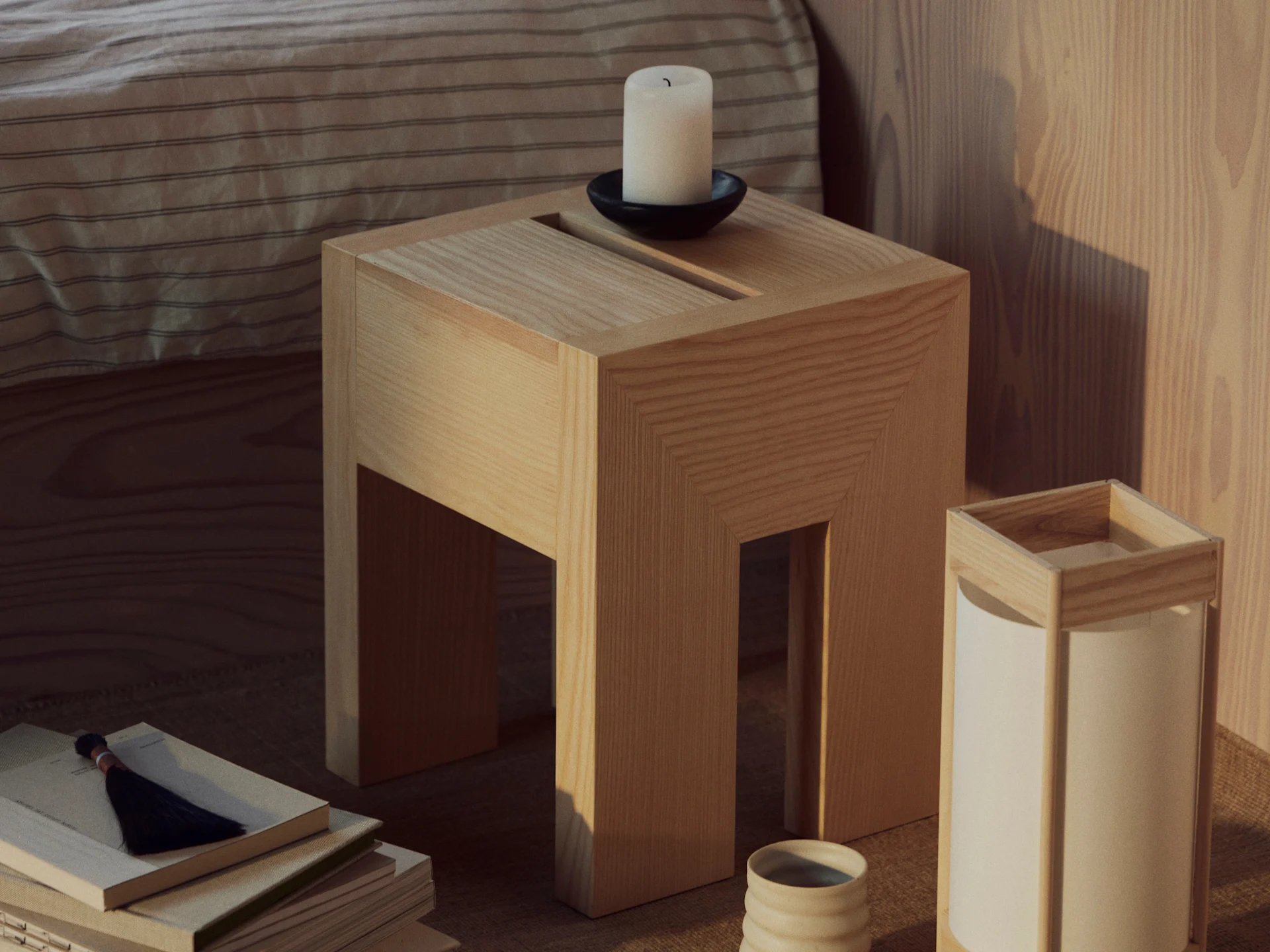 Una mesa auxiliar de madera con una vela blanca en un soporte negro, una pila de libros con borla y una lámpara con taza cerámica.