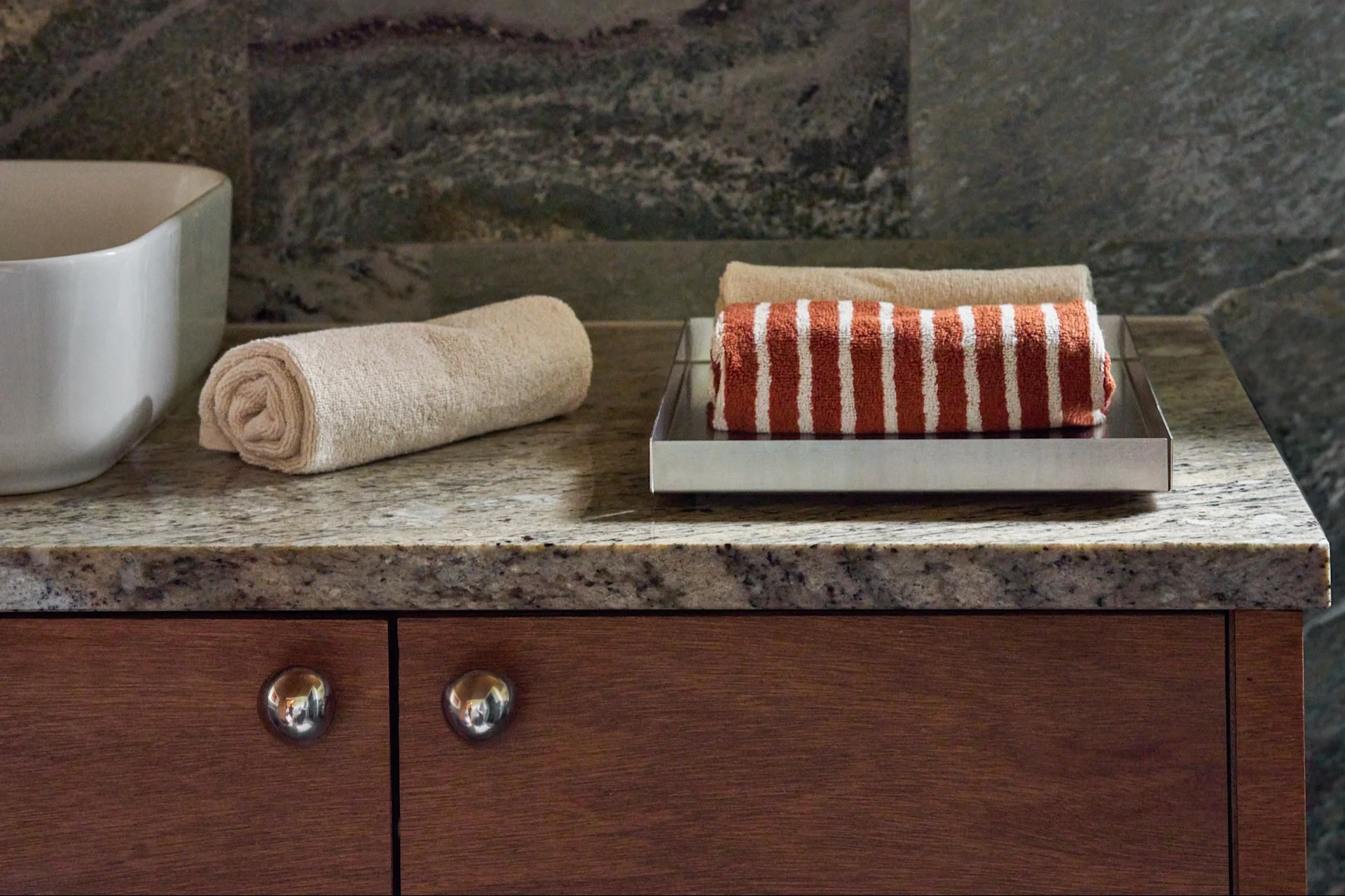 Toallas enrolladas de color beige y rayas rojizas sobre una bandeja plateada en una encimera de granito, junto a un lavabo blanco y gabinetes de madera.