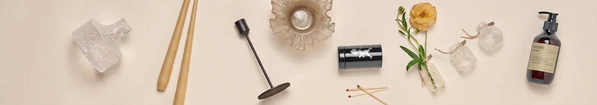 Una colección de artículos de decoración para el hogar, incluyendo jarrones de vidrio, palillos, un candelabro negro, un cuenco de vidrio con volantes, un recipiente negro, una flor naranja y una botella de loción de manos.