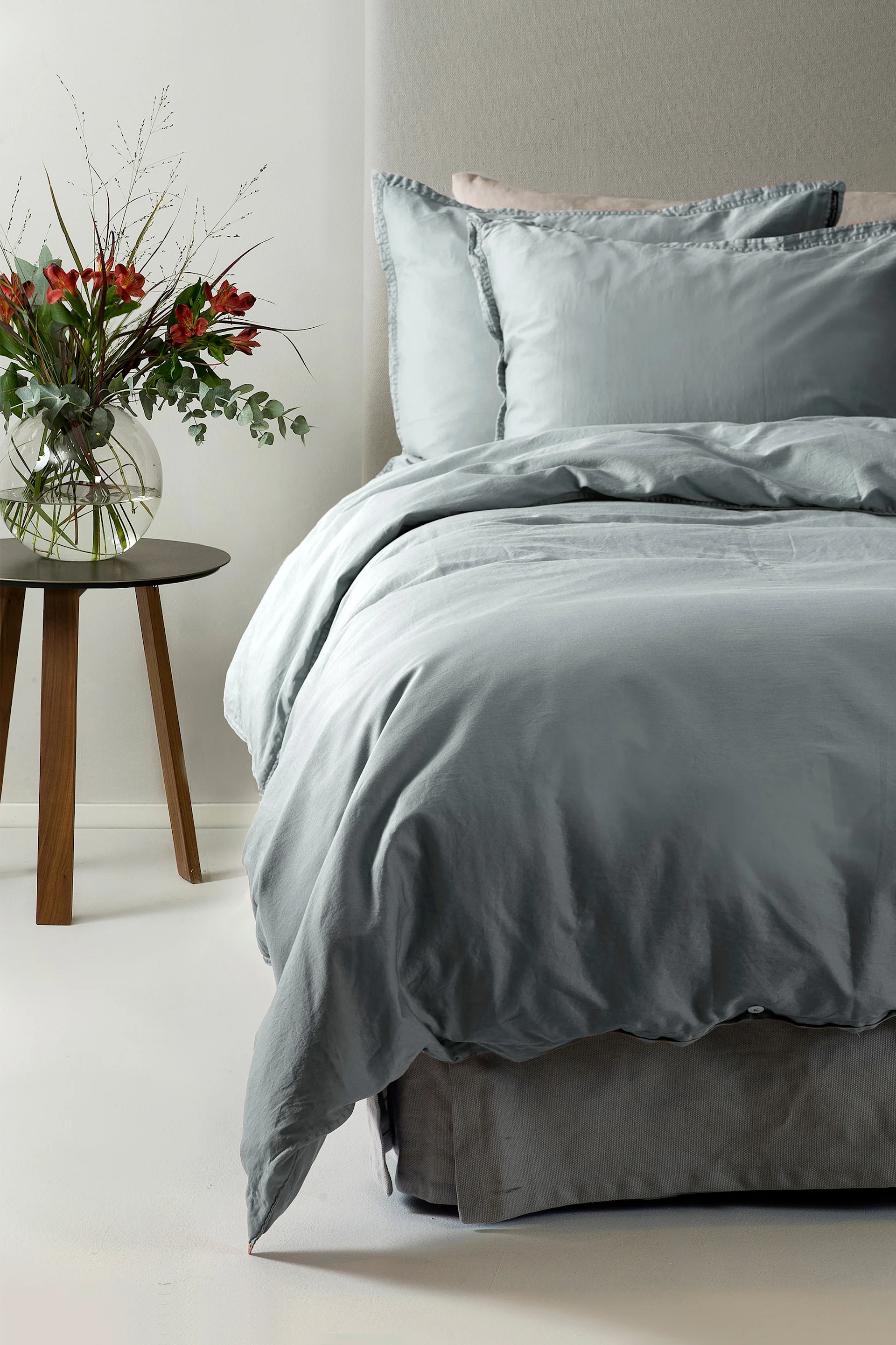 Una cama con ropa de cama azul claro y dos almohadas, junto a una mesita de madera con un jarrón de flores rojas.