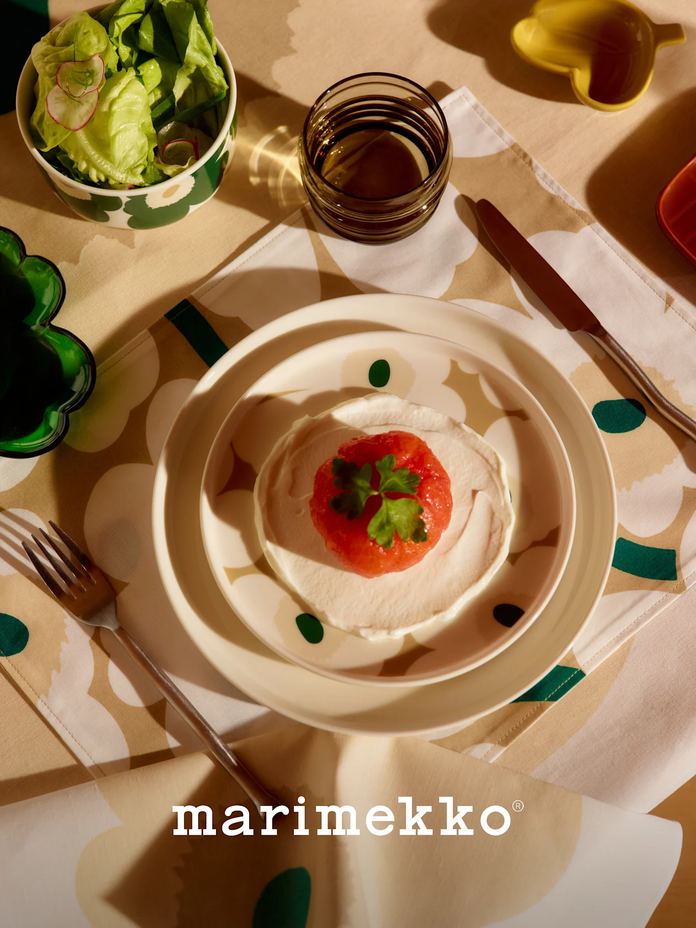 Mesa Marimekko con cuenco de ensalada estampado, plato de fruta roja y crema, vaso y cubiertos sobre mantel floral.