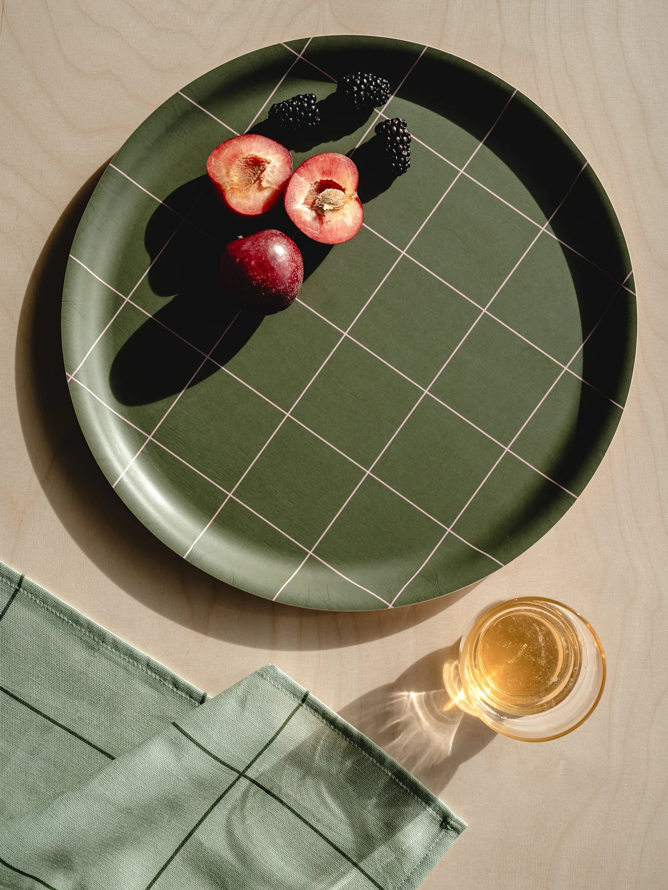 Bandeja verde oscuro a cuadros con ciruelas rojas, moras, servilleta verde claro y un vaso de zumo sobre madera.
