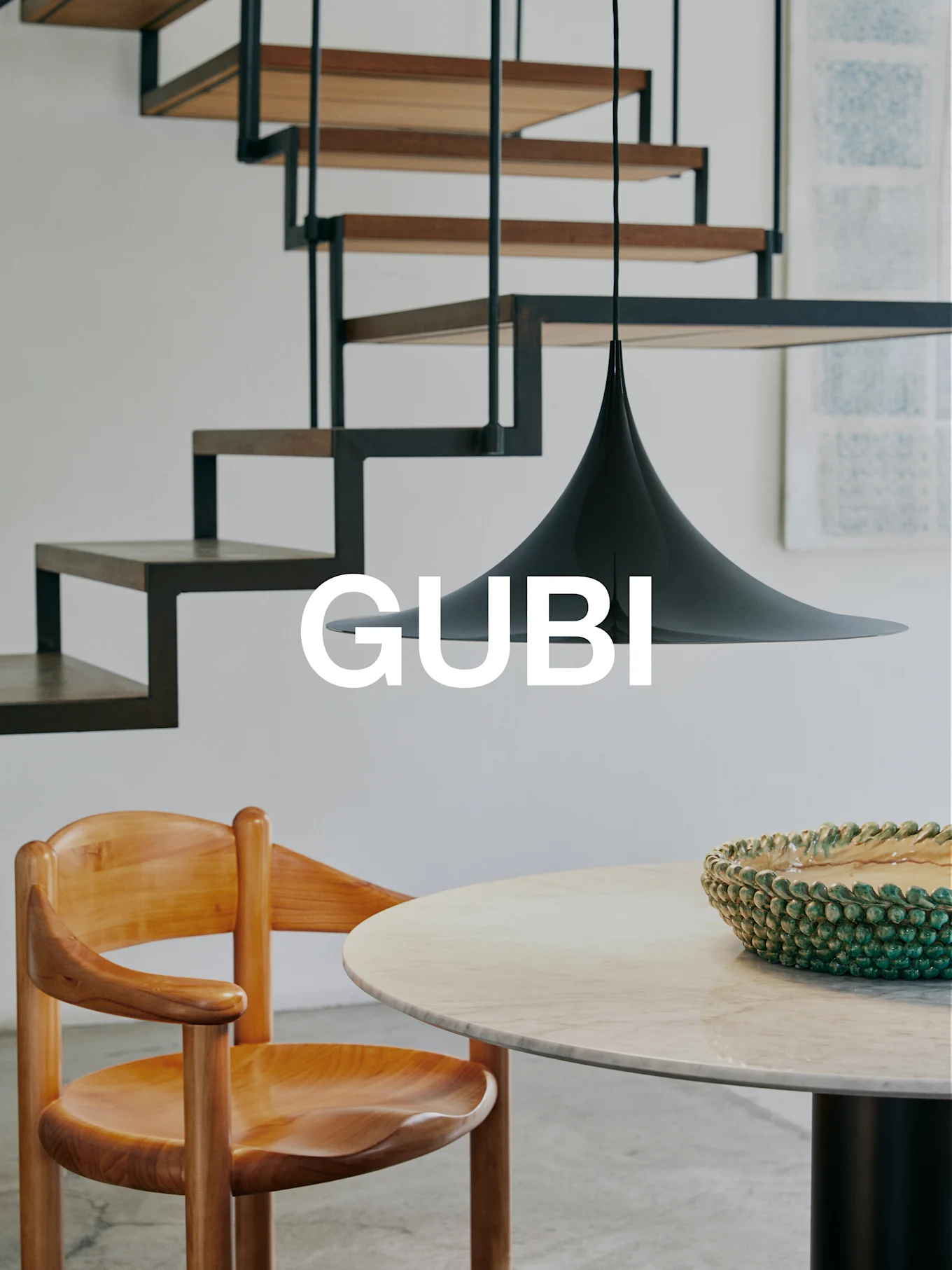 Logotipo GUBI en un interior moderno con silla de madera, mesa de mármol, cuenco verde, lámpara colgante negra y escalera moderna.