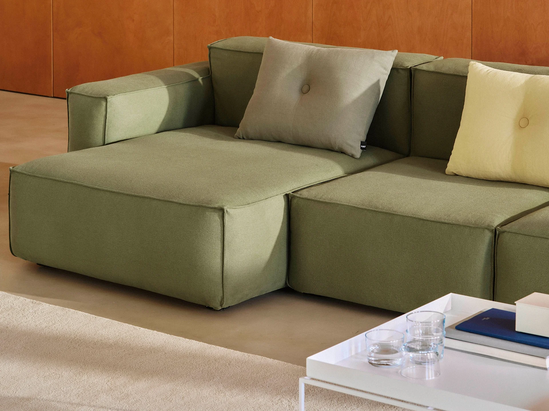 Un sofá modular verde oliva con cojines decorativos gris claro y amarillo, junto a una mesa de centro blanca con libros.
