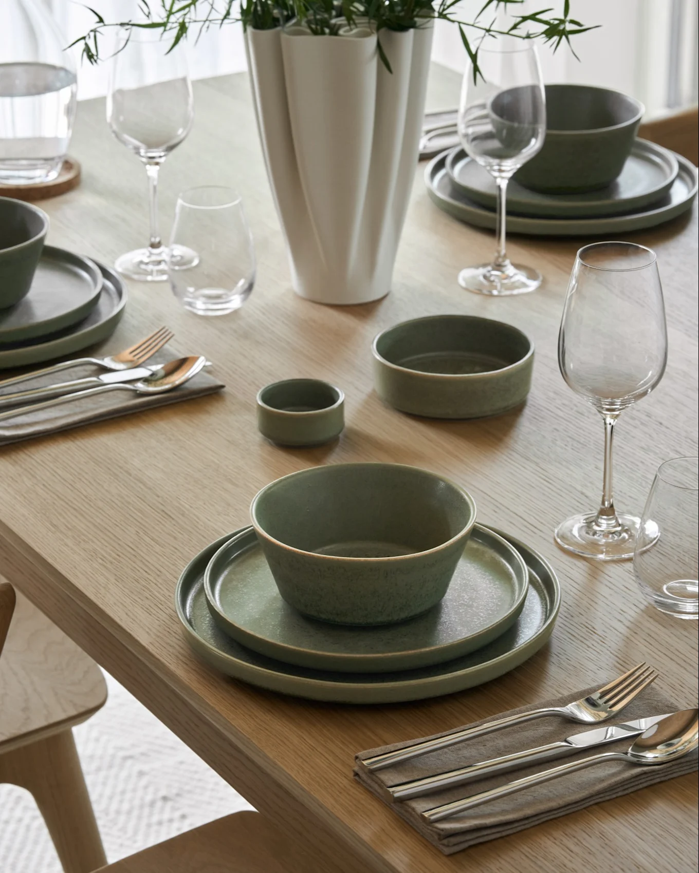 Mesa de comedor puesta con cuencos de sopa cremosa, platos a juego, copas de vino y agua, cubiertos y un jarrón con follaje verde.