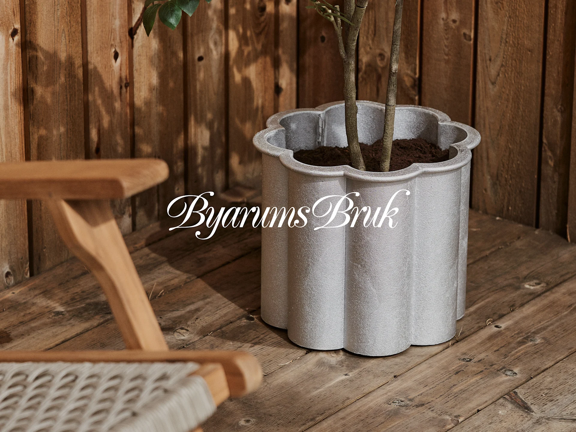 Una maceta festoneada gris plateado con una planta en una terraza de madera, junto a una silla de madera. Logotipo 'Byarums Bruk'.