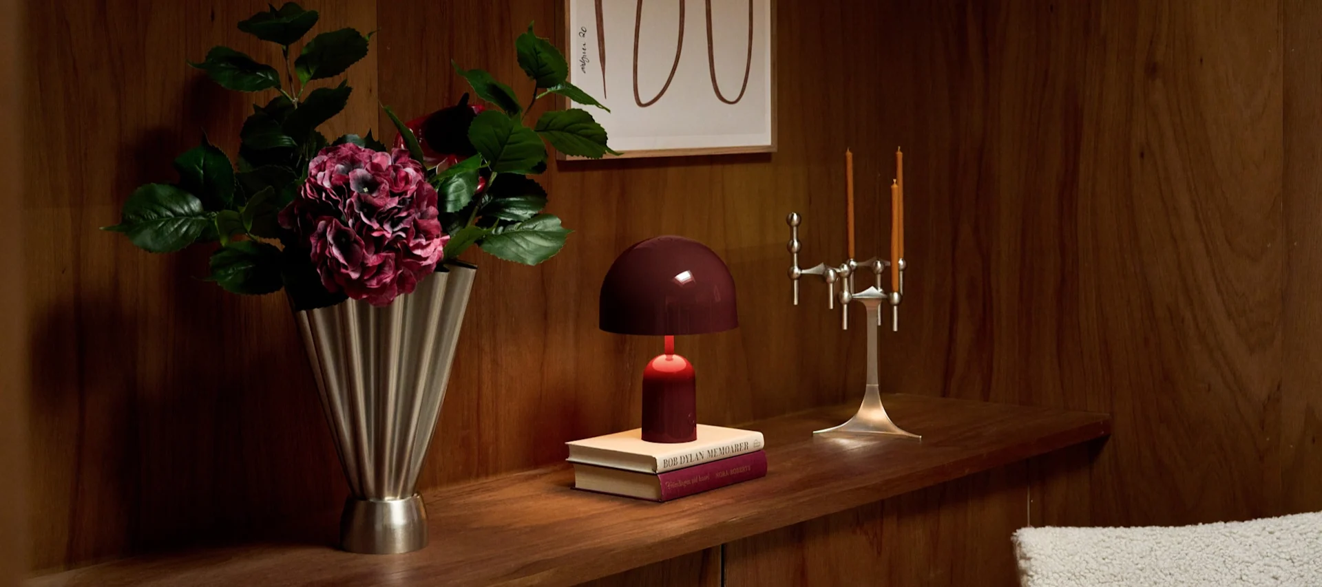 Estante de madera: jarrón plateado con hortensias oscuras, lámpara burdeos en libros, candelabro plateado con velas naranjas.