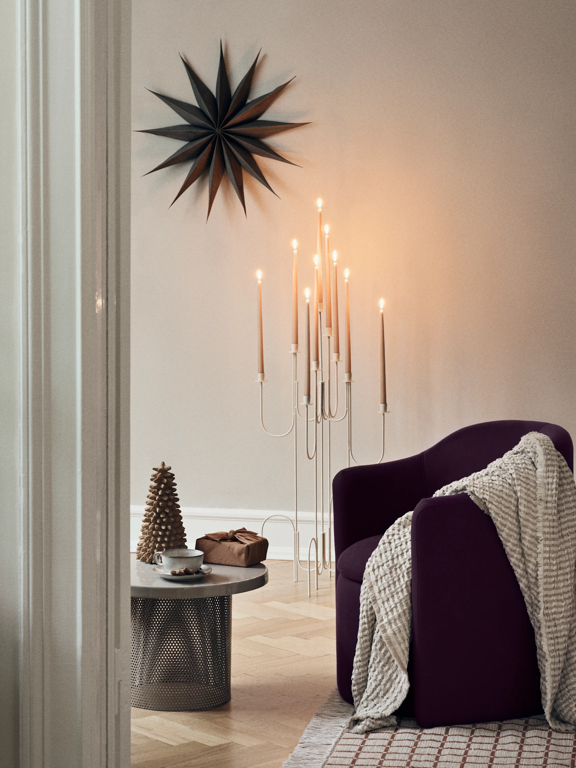Una escena invernal luminosa a través de una ventana, con árboles nevados y un estallido de sol. Dentro, una mesa con velas y árboles de papel, y una estrella de papel cuelga en la ventana.