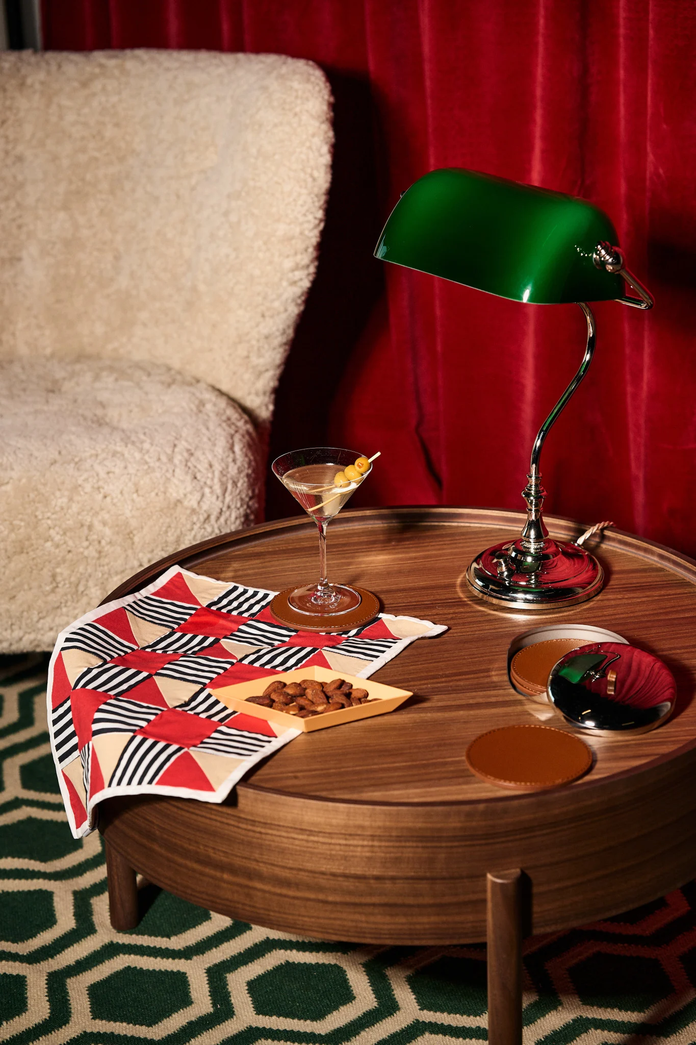 Martini, almendras, lámpara de banquero verde en mesa de madera con tela geométrica, sillón crema y cortinas rojas.