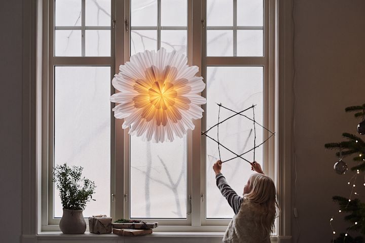 Estrella de adviento Snöblomma de Watt & Veke en una ventana, con una niña al lado sosteniendo una estrella hecha de ramas.