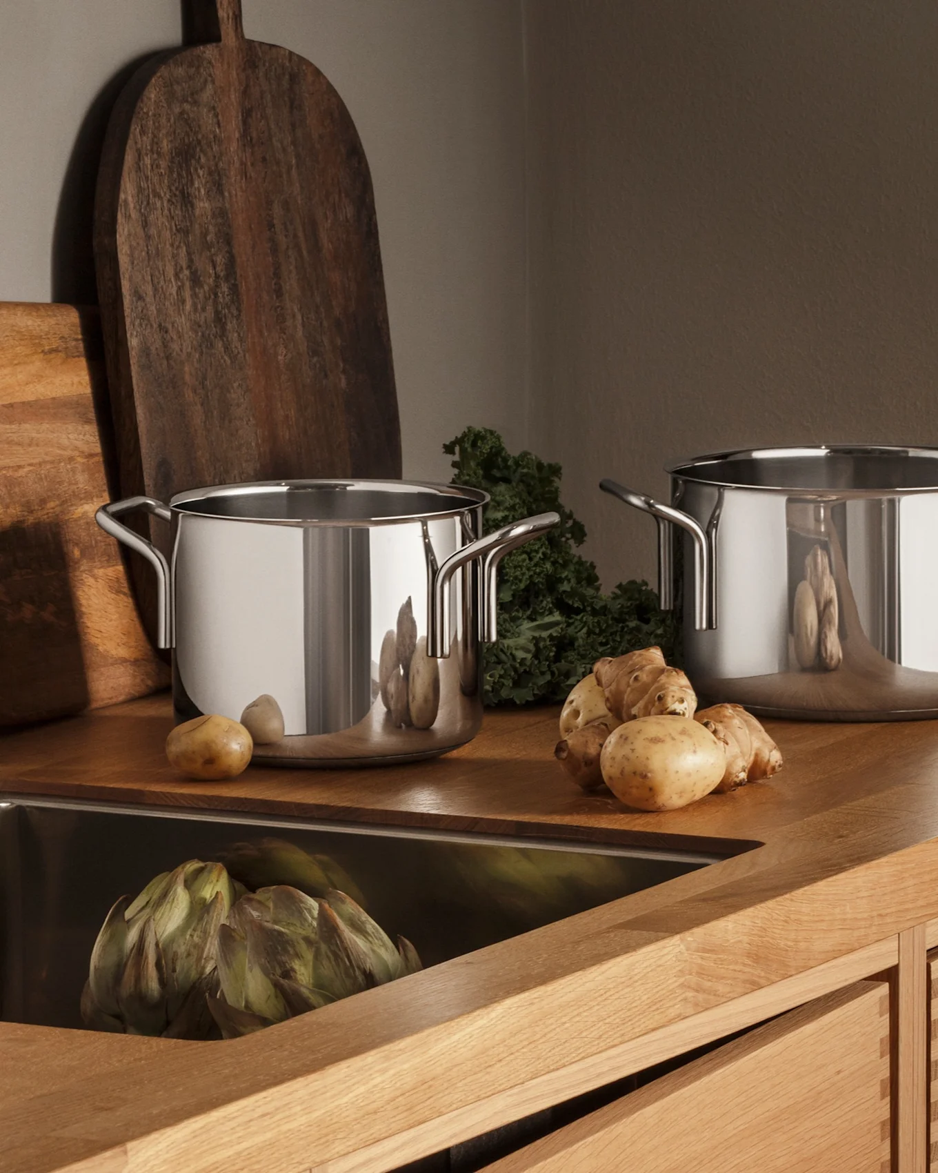 Cocina moderna con encimera de madera, fregadero de acero inoxidable, grifo, tres ollas y productos frescos como patatas y alcachofas.
