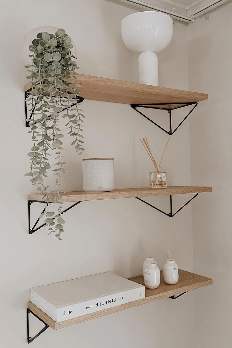 Tres estantes flotantes de madera clara con soportes triangulares negros. Los artículos incluyen una planta colgante, un jarrón blanco, un recipiente con puntos, un difusor, un libro blanco y jarrones pequeños blancos.