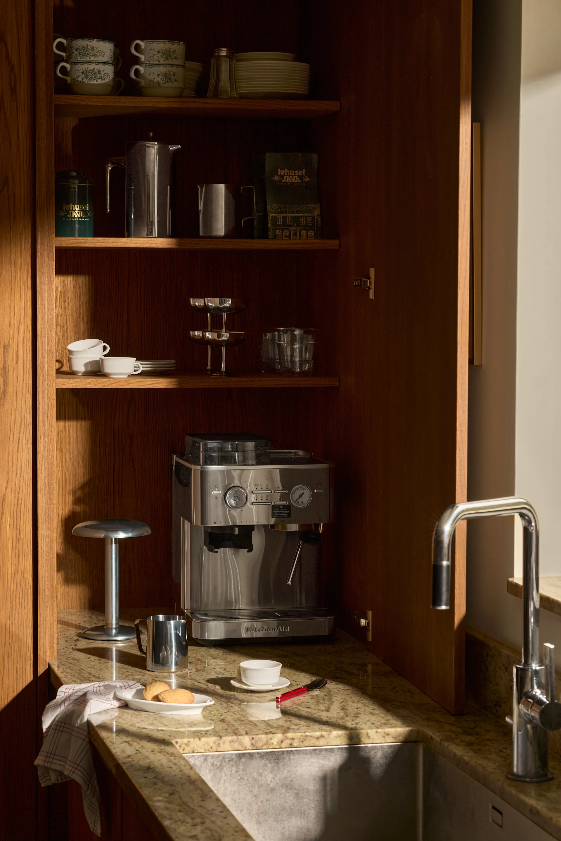 Armario de madera abierto con cafetera espresso KitchenAid, tazas, platos, artículos de café y fregadero en encimera de granito.