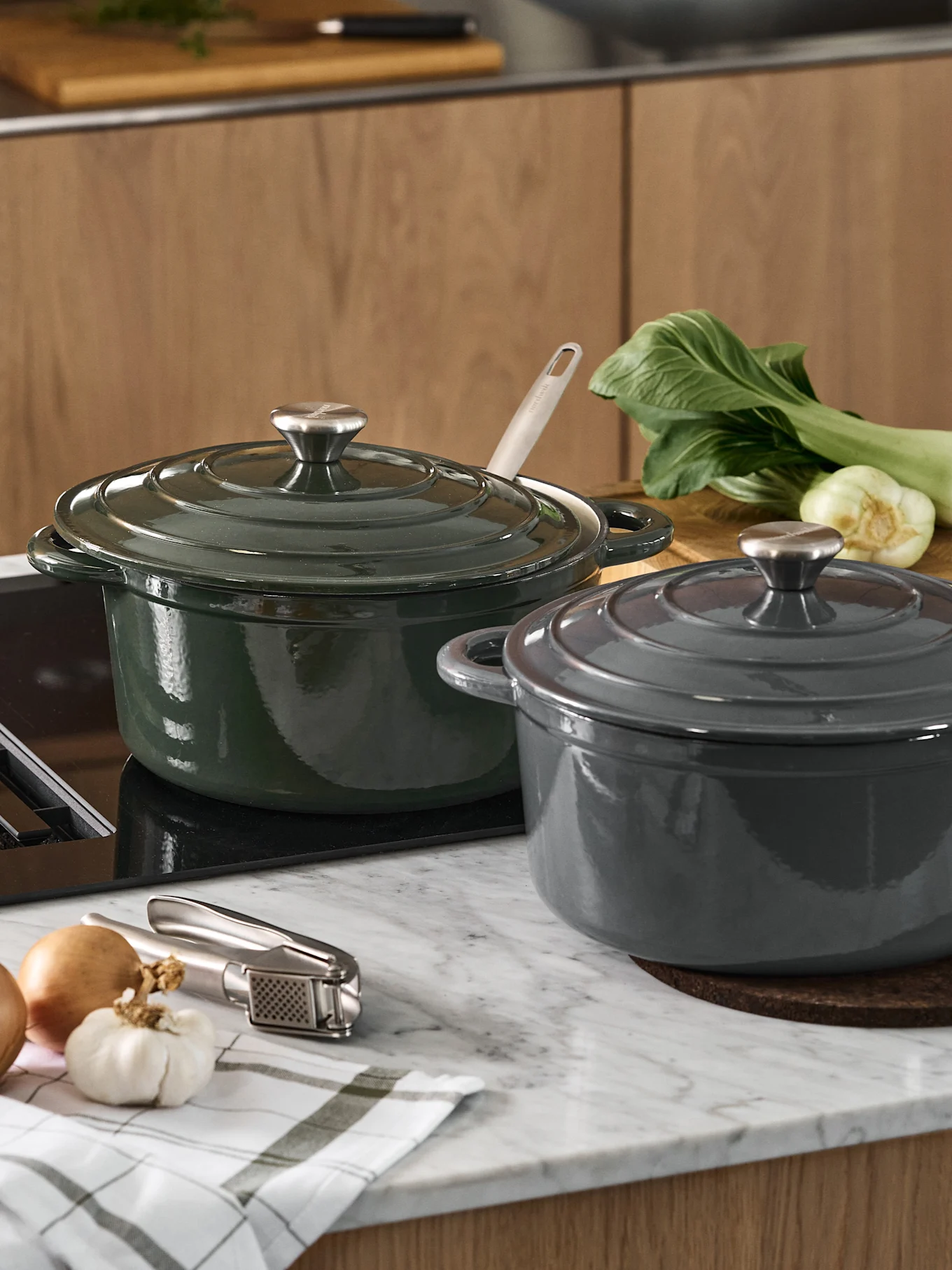 Dos ollas de hierro fundido con tapa, una verde oscuro y otra gris oscuro, sobre una encimera de cocina con verduras y un prensador de ajos.