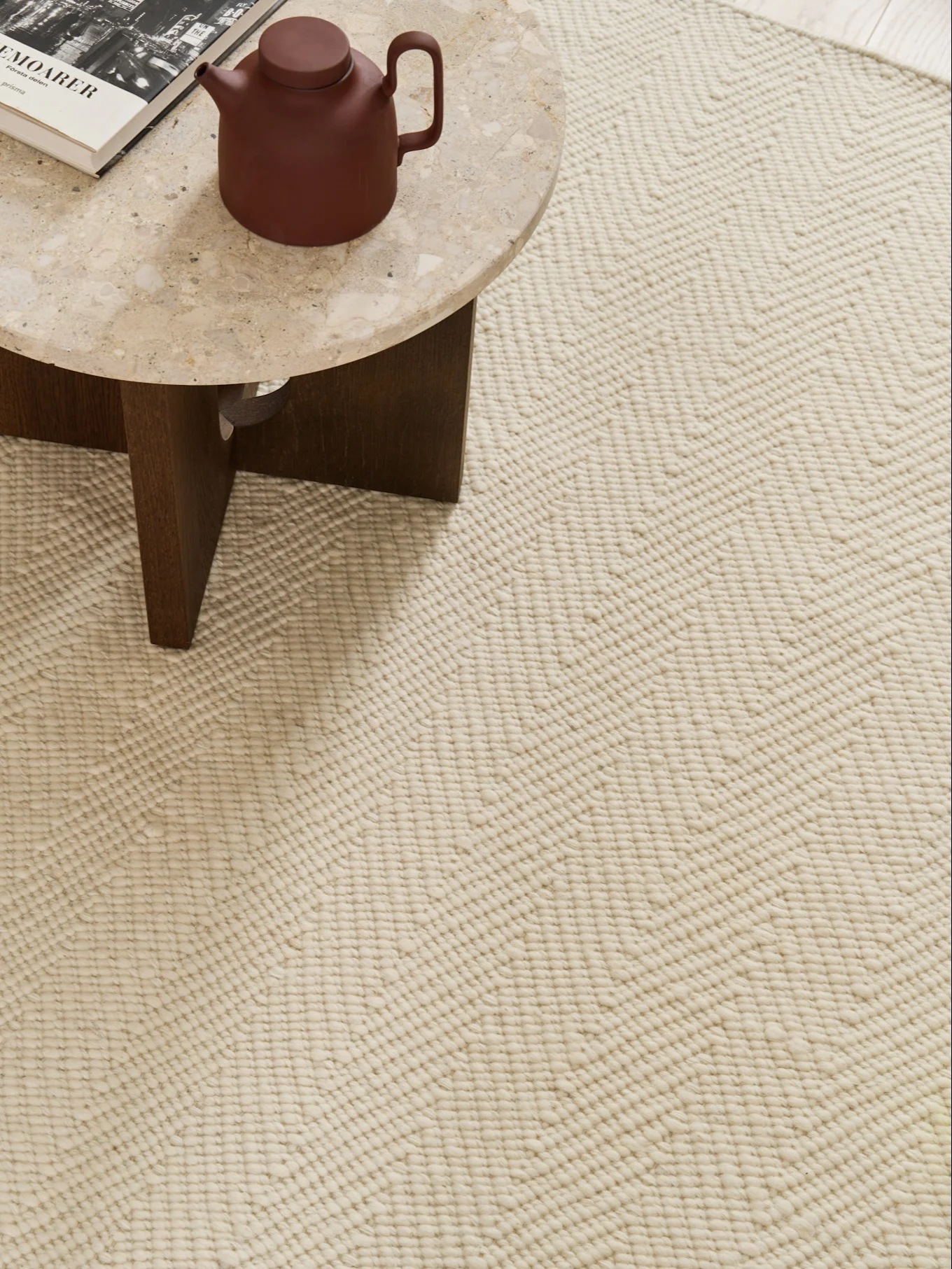 Primer plano de una alfombra crema con patrón de zigzag texturizado, con una mesa de mármol con tetera y libro en la esquina.