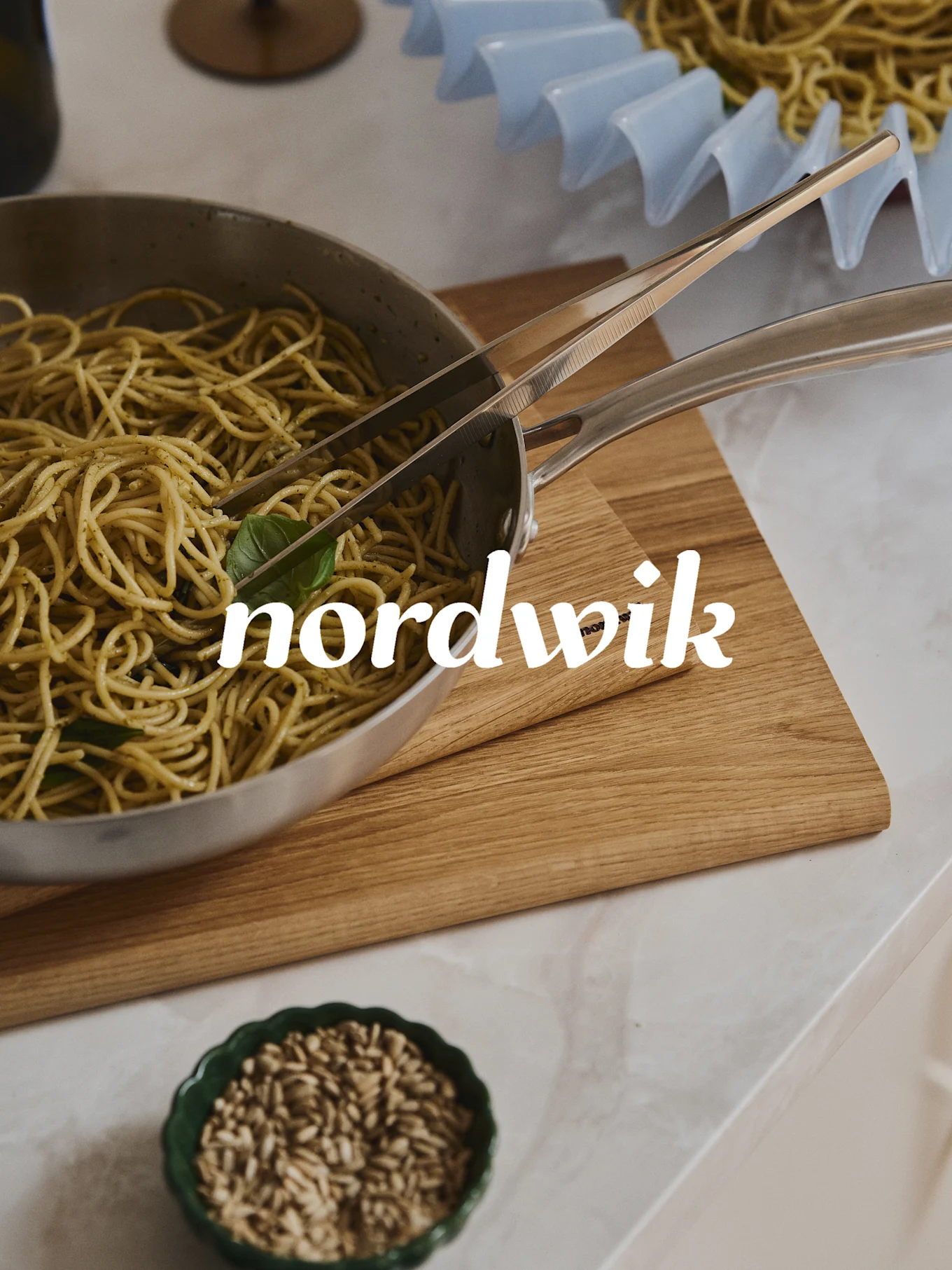 Espaguetis con pesto en sartén de acero con pinzas sobre tabla de madera. Un cuenco de semillas debajo, y un escurridor de pasta al fondo. Superposición de texto 'nordwik'.