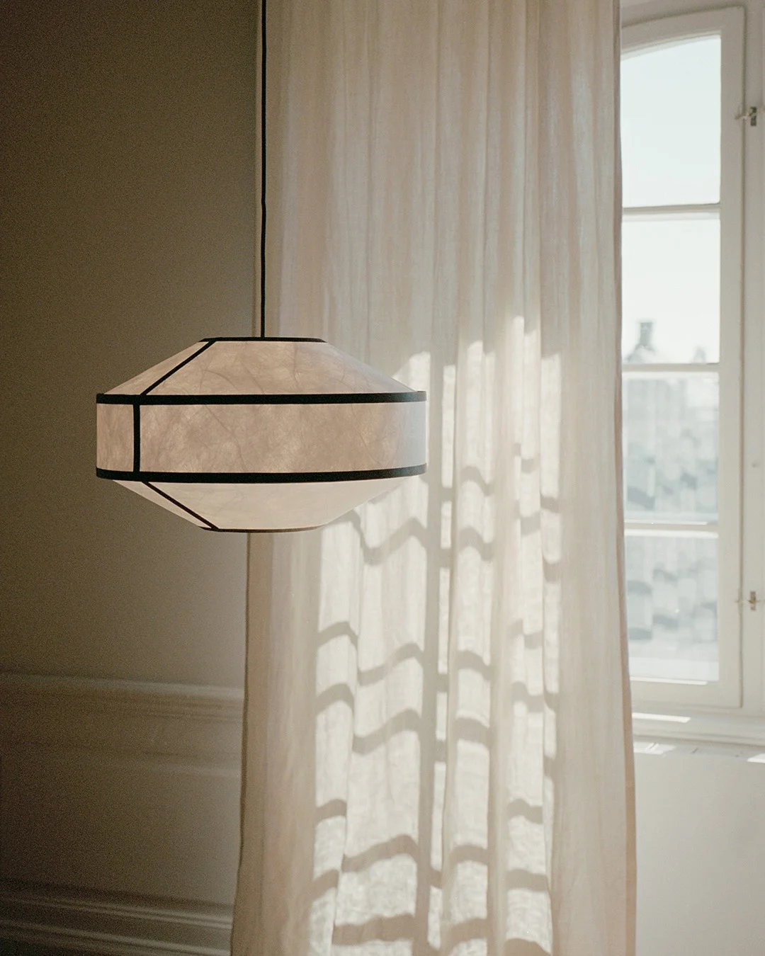Una lámpara colgante geométrica de papel con marcos oscuros cuelga junto a una cortina translúcida y ventana iluminadas por el sol.