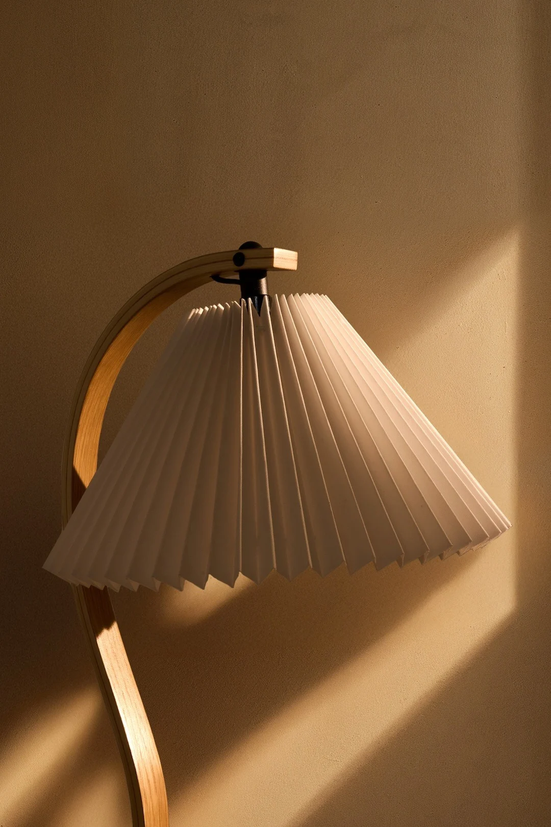 Una lámpara con una pantalla blanca plisada y una base de madera curvada, proyectando luz cálida sobre una pared texturizada.