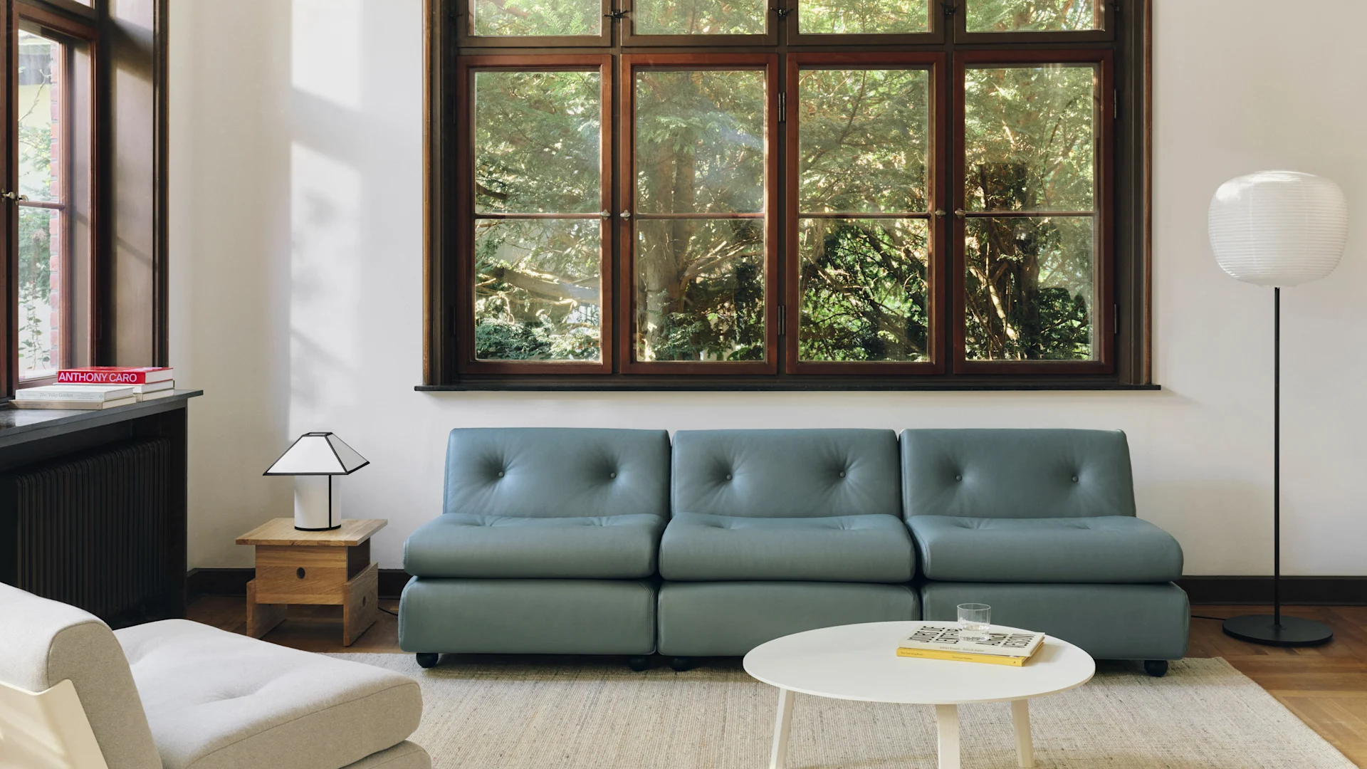 Sala de estar moderna con un sofá modular azul grisáceo, mesa de café blanca y una gran ventana con vistas a árboles verdes. Dos lámparas iluminan el espacio.