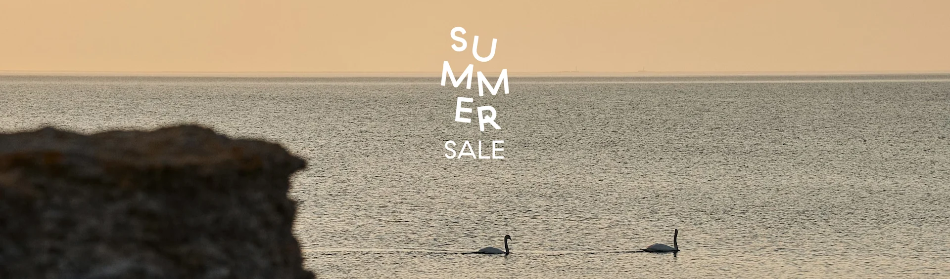 Dos cisnes nadan en aguas tranquilas bajo un cielo cálido con una roca oscura en primer plano y texto 'SUMMER SALE'.