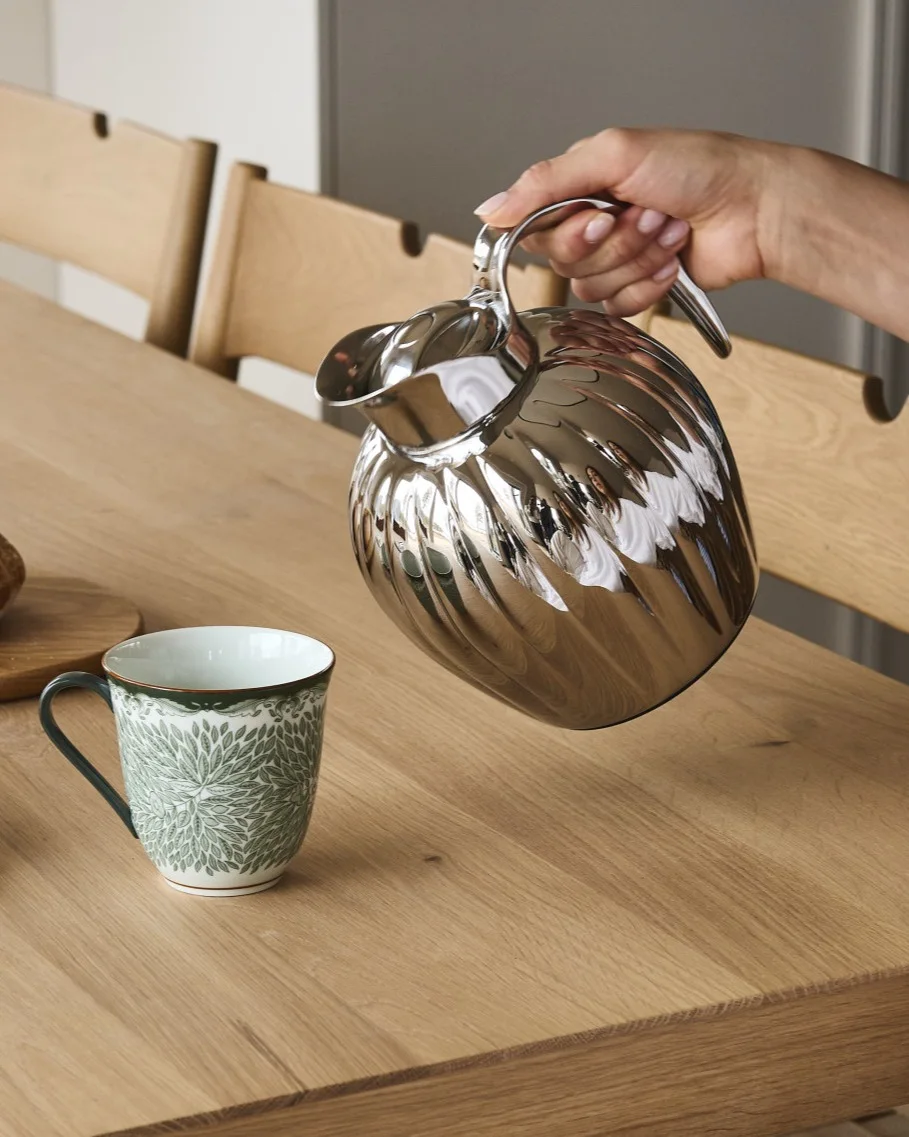 Una mano vierte de una jarra estriada plateada en una taza blanca con un patrón de hojas verdes sobre una mesa de madera.