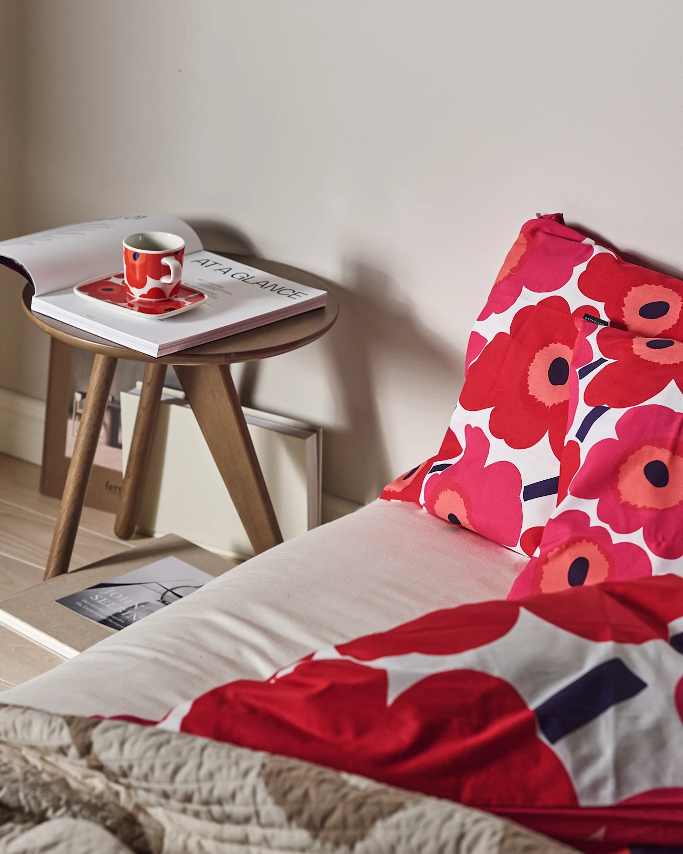 Una cama con ropa de cama floral roja y rosa, y una manta beige texturizada. Una taza roja y un libro abierto en una mesita de madera.