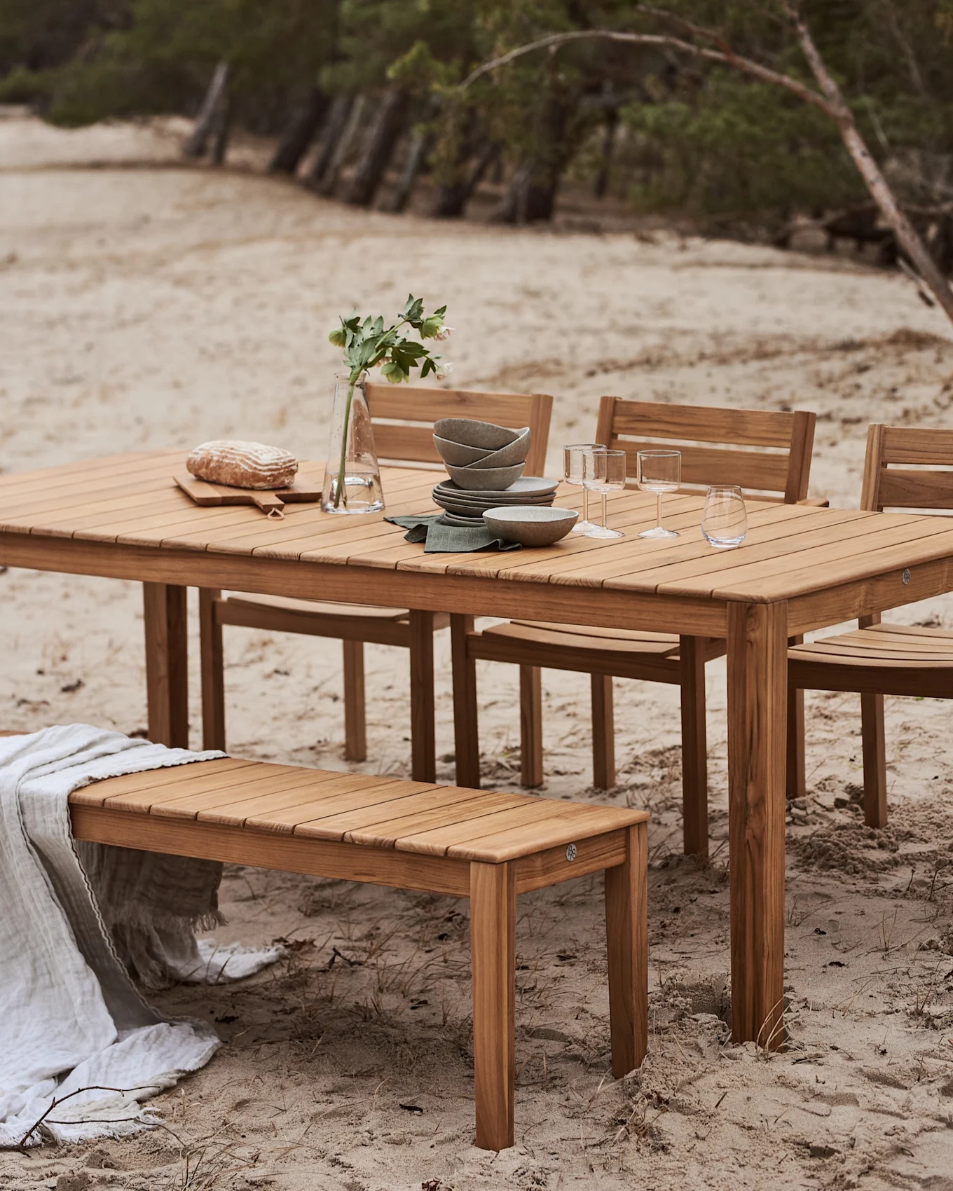 Juego de comedor de madera para exterior con mesa y banco en una playa de arena. La mesa está puesta con vajilla, vasos, pan y un jarrón con flores.