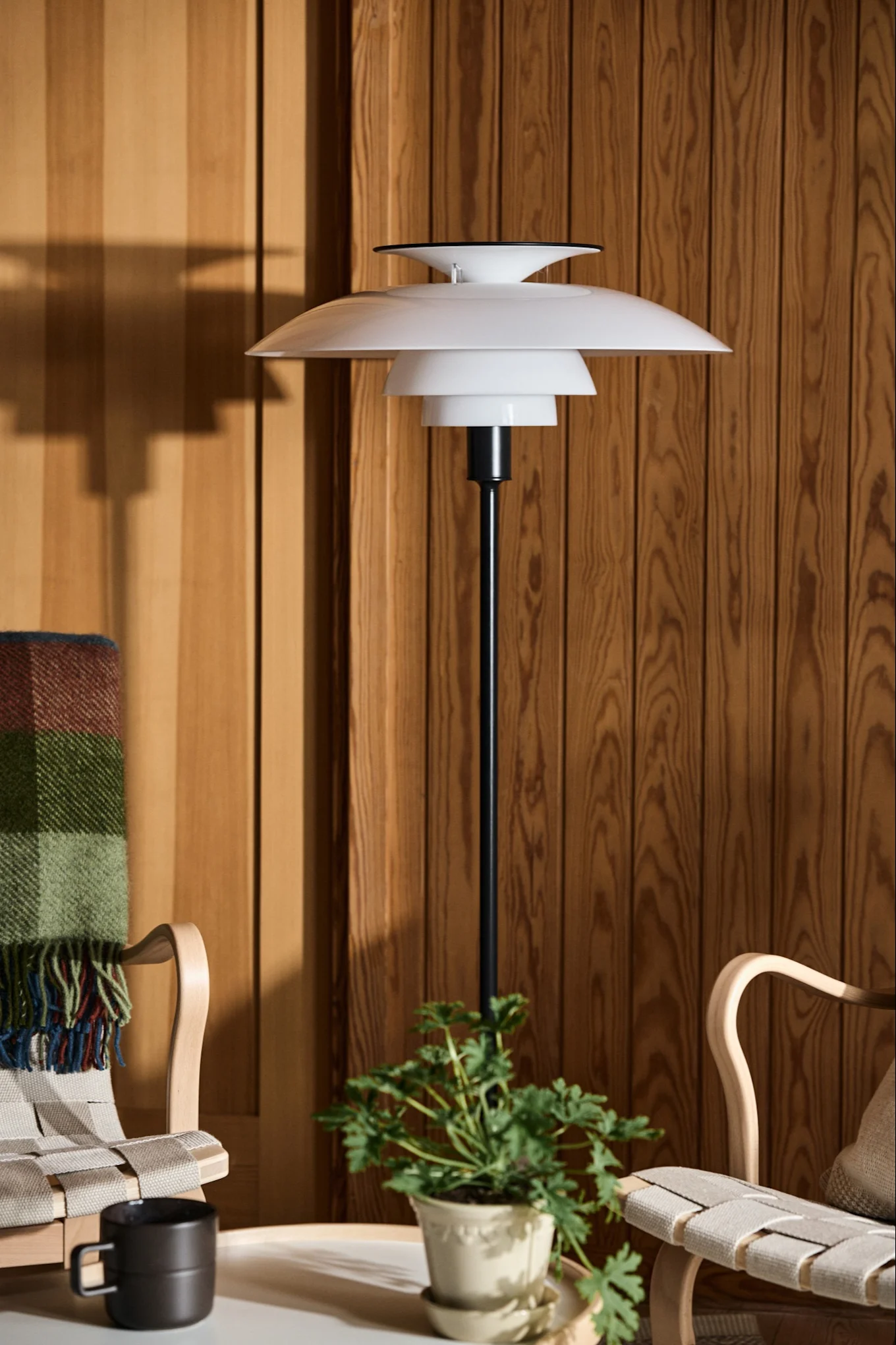 Lámpara de pie blanca con capas junto a dos sillas de madera y una planta contra una pared de madera.