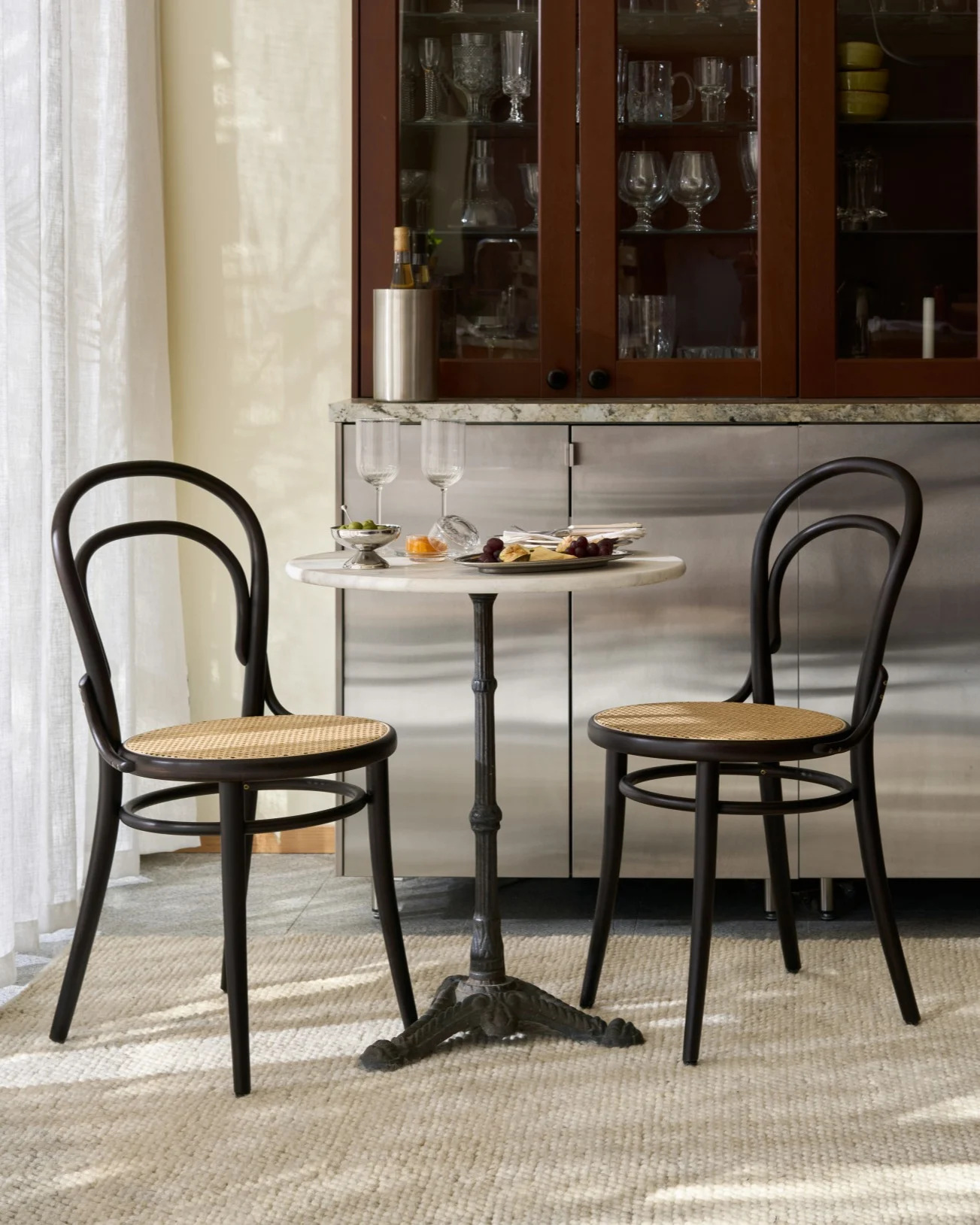 Una mesa de bistró con dos sillas de madera y caña, con aperitivos y copas, frente a un armario con cristalería.