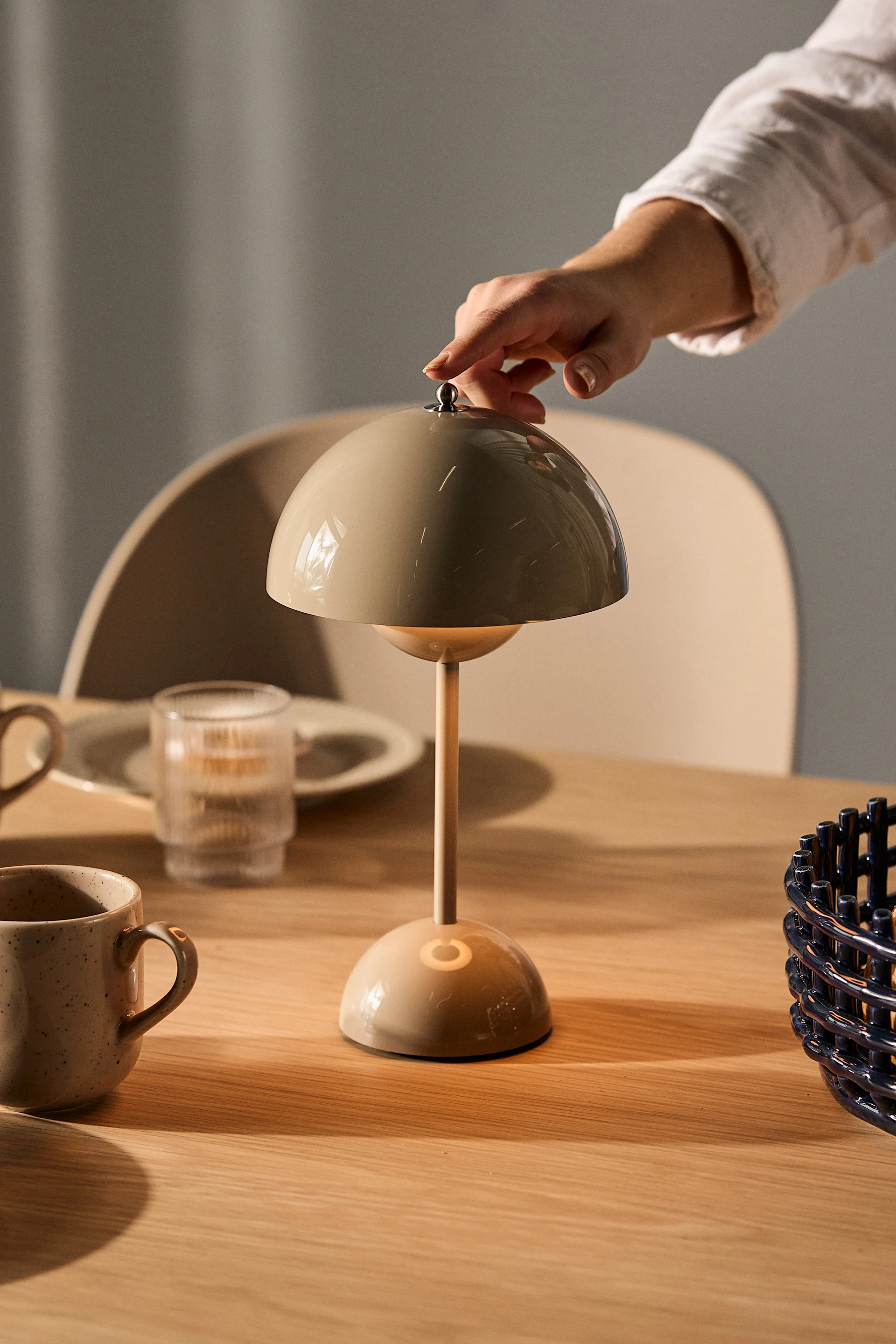 Una mano ajusta una lámpara de mesa beige con forma de hongo sobre una mesa de madera con tazas y un vaso.