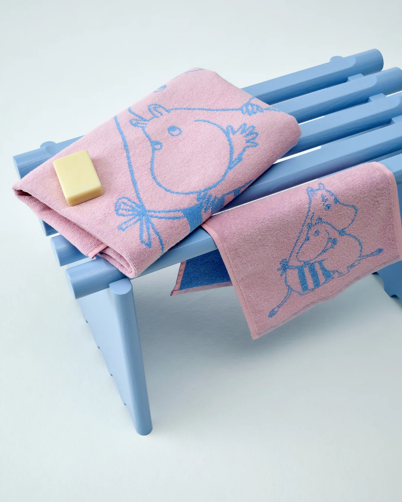 Dos toallas Moomin rosas con estampados azules, una doblada con una barra de jabón, sobre un banco azul claro.