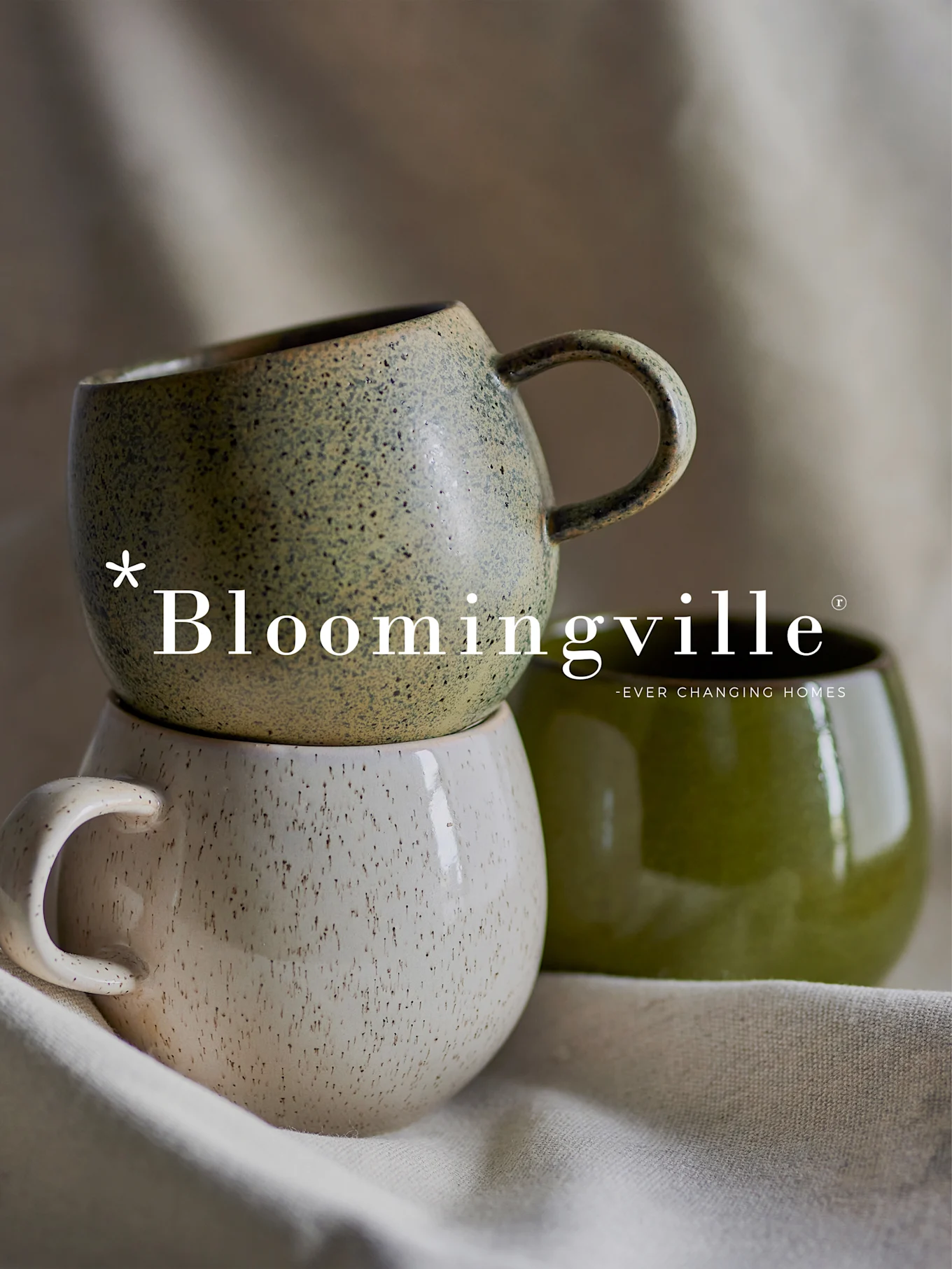 Logotipo de Bloomingville sobre una pila de dos tazas de cerámica moteadas en verde y blanco roto, con otra taza verde lisa a su lado.