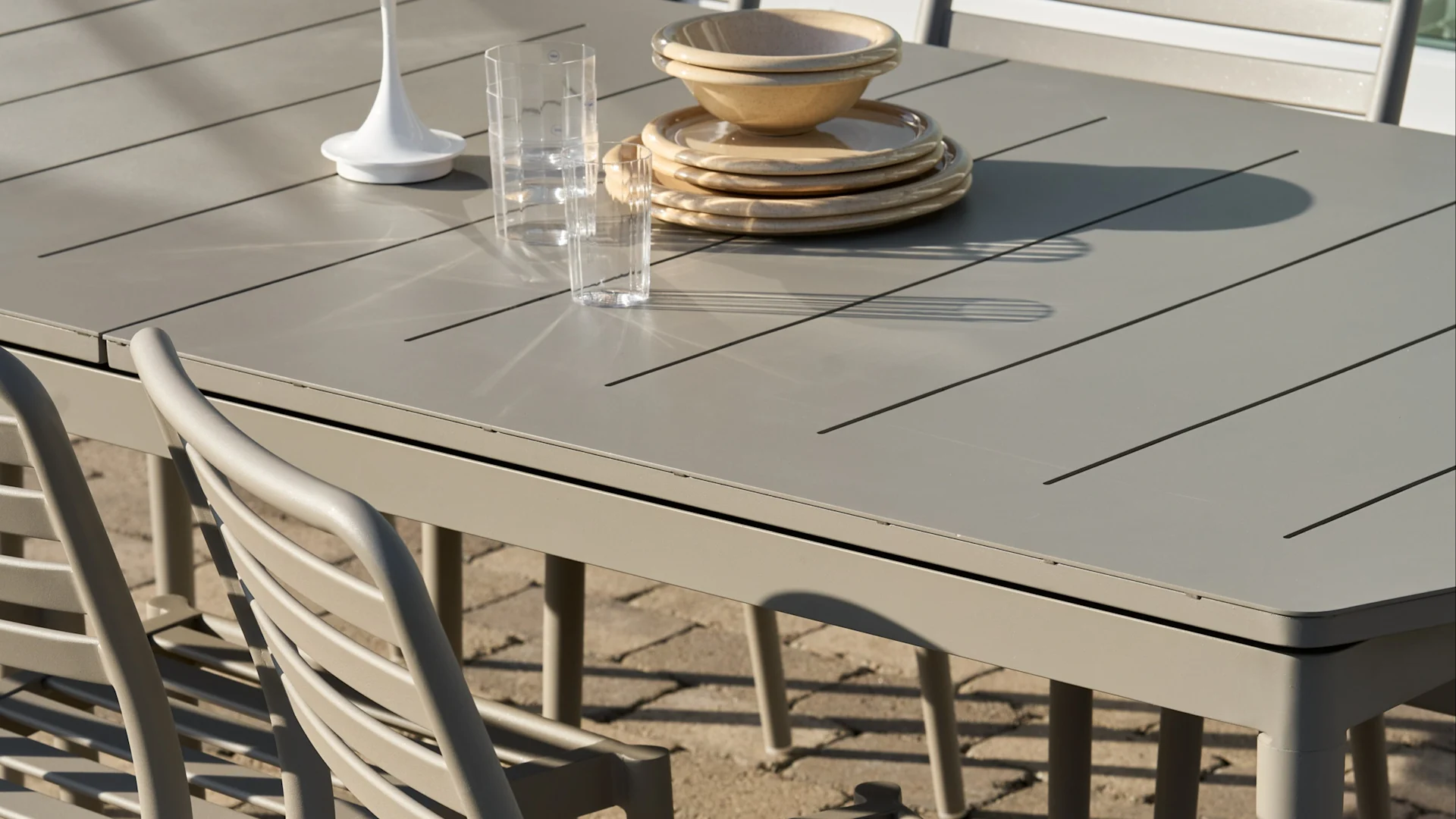 Mesa de comedor exterior de lamas gris claro con platos y cuencos beige apilados, dos vasos transparentes y un adorno blanco.