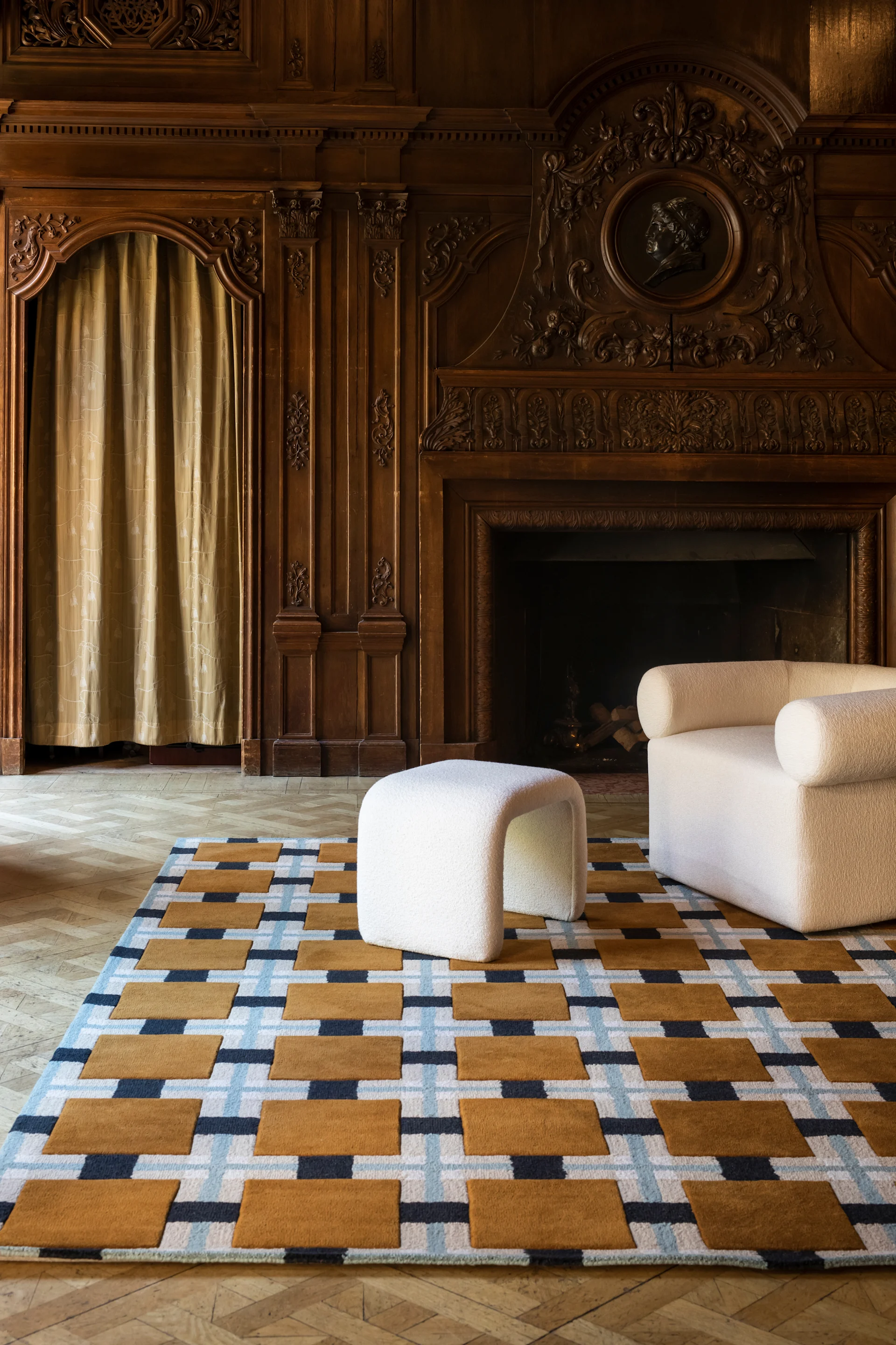 Alfombra geométrica con patrones marrones, azules y blancos, un puf y sillón blancos en una habitación con paneles de madera.