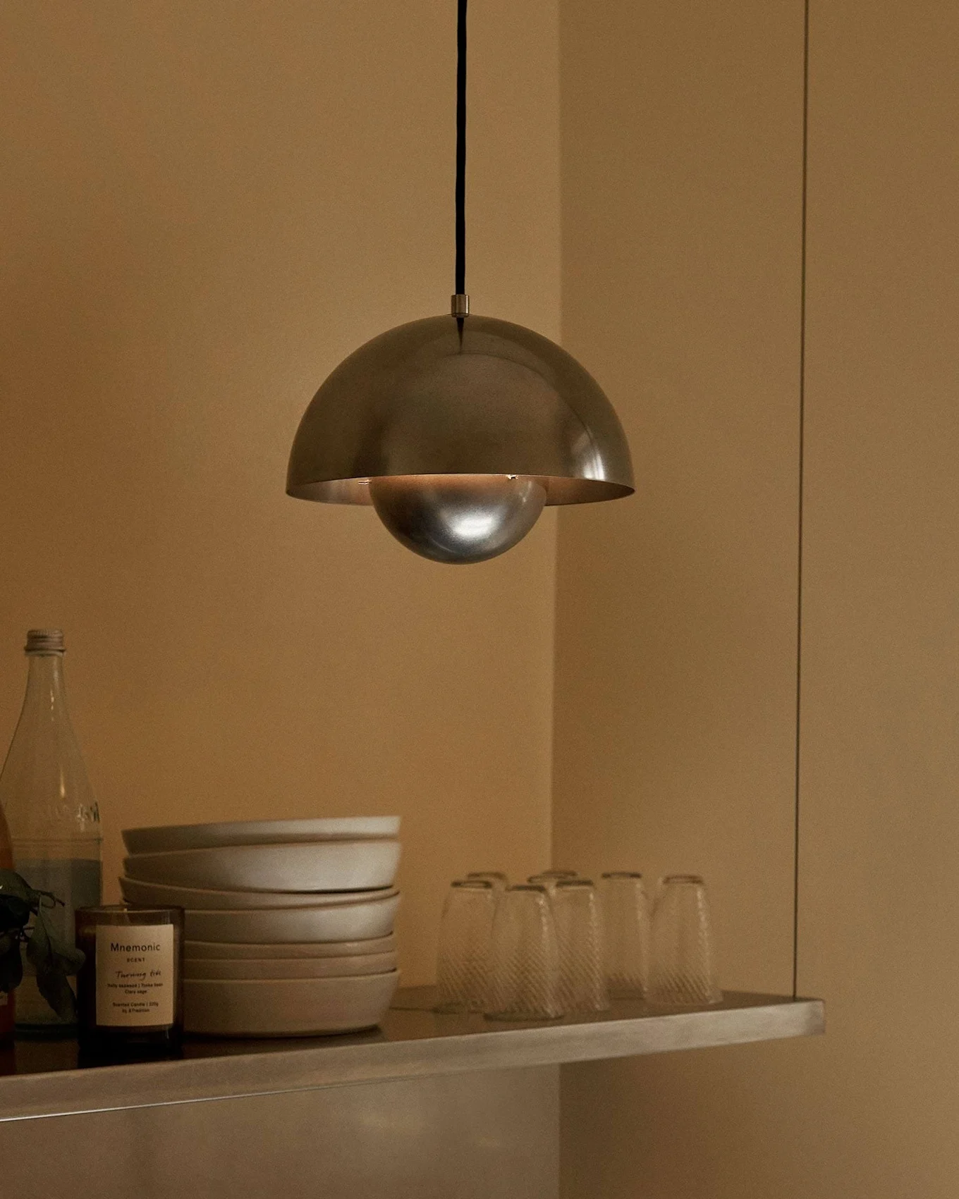 Una lámpara colgante de metal en forma de cúpula cuelga sobre un estante con cuencos, vasos, botellas, una fruta y una vela.