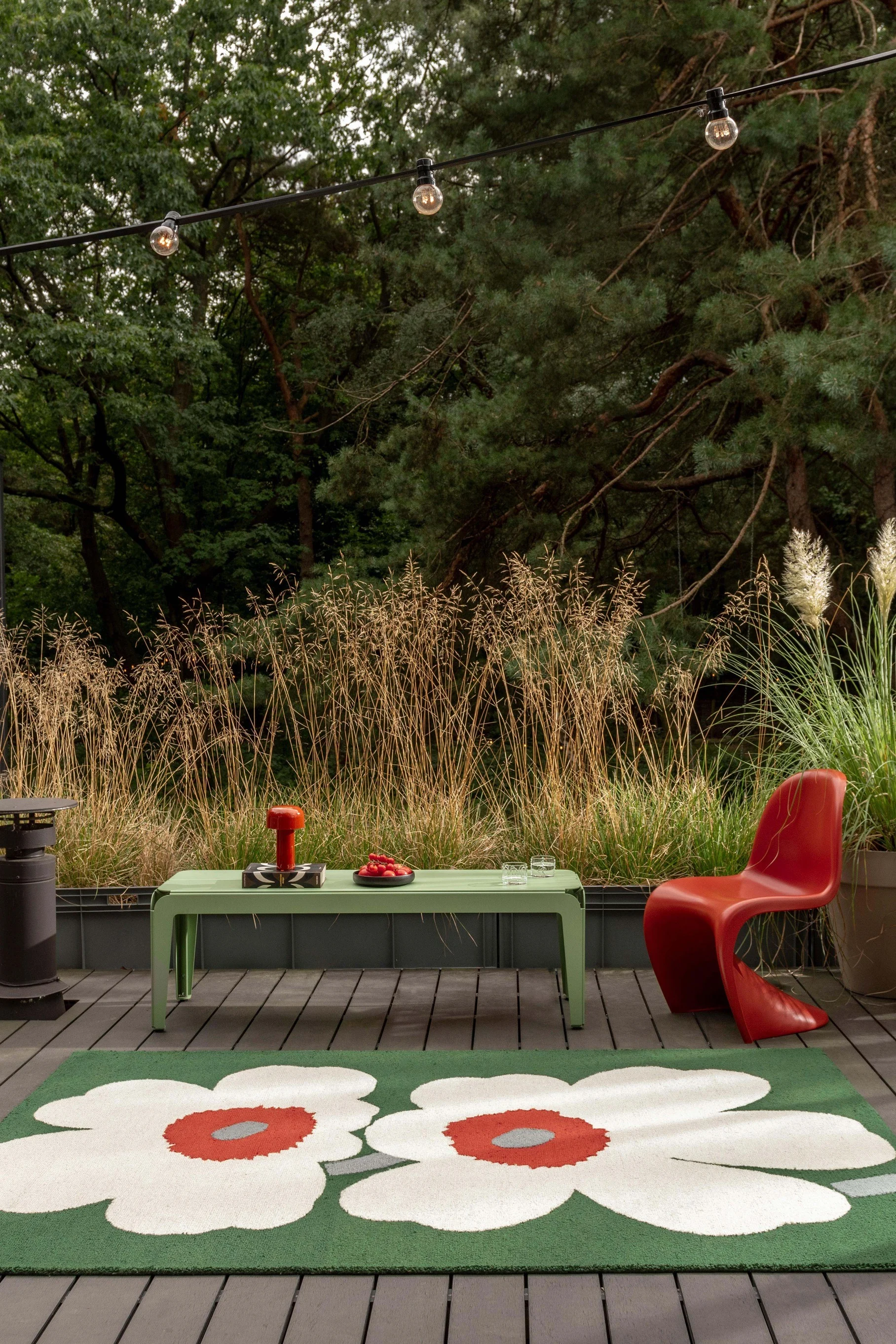 Un patio exterior con una alfombra verde floral, una mesa verde claro, una silla roja y luces de cadena, rodeado de hierbas altas y árboles.