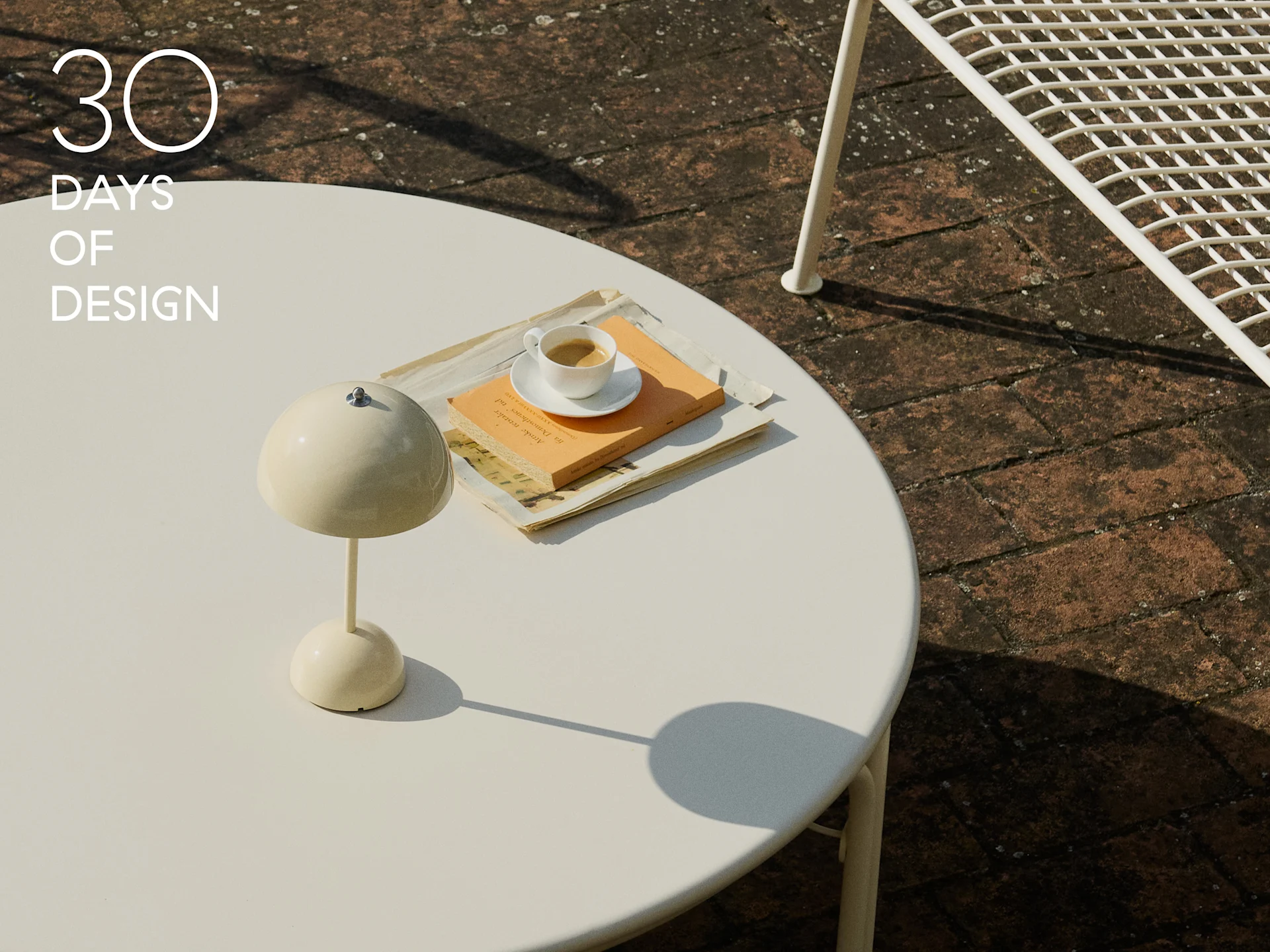 Lámpara de seta crema, taza de café y libro sobre una mesa redonda blanca de exterior.