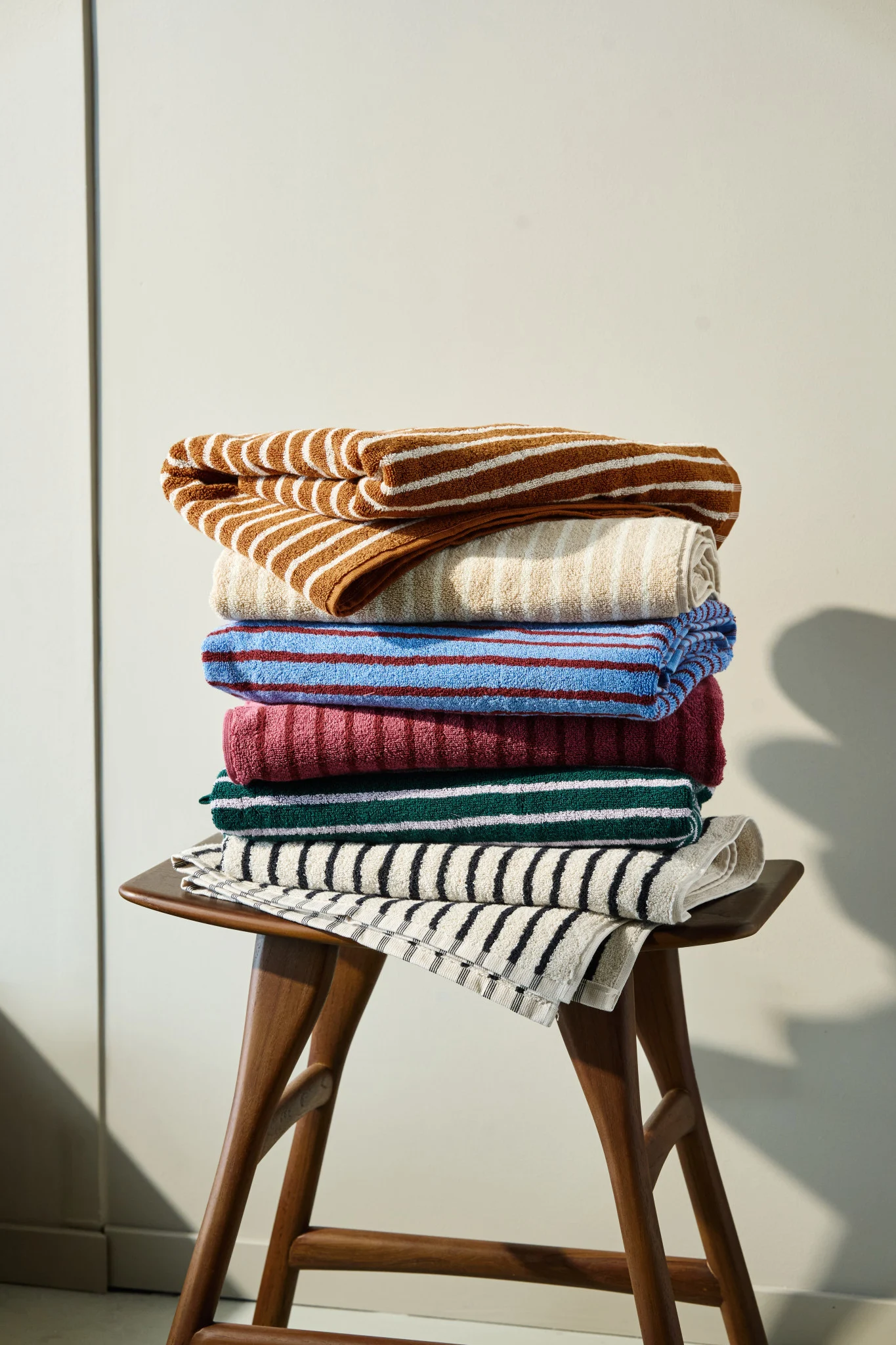 Una pila de toallas de rayas y lisas de colores marrón, beige, azul, granate, verde y negro sobre un taburete de madera.