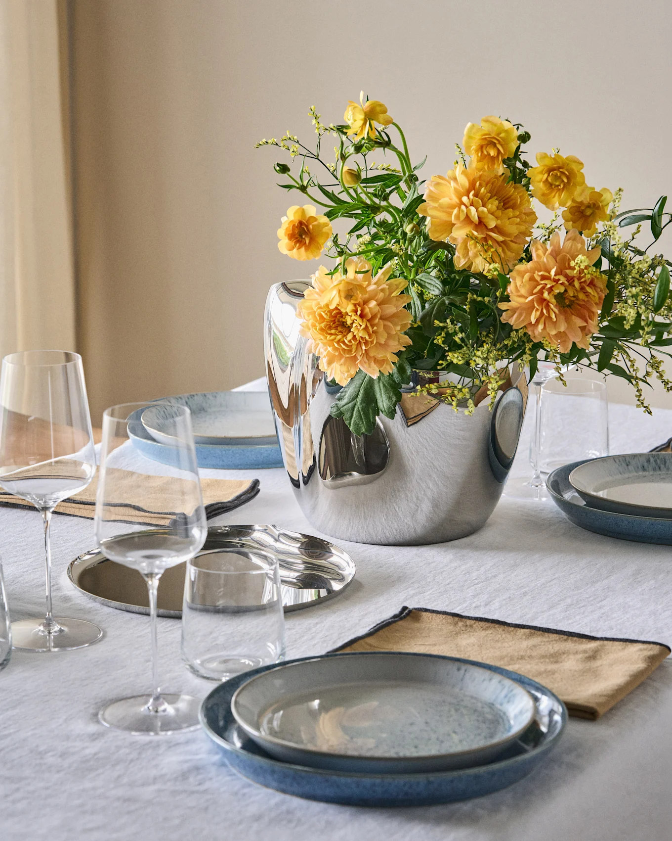 Una mesa de comedor puesta con platos azules, servilletas beige, vasos transparentes y un jarrón plateado con flores naranjas y amarillas sobre un mantel blanco.