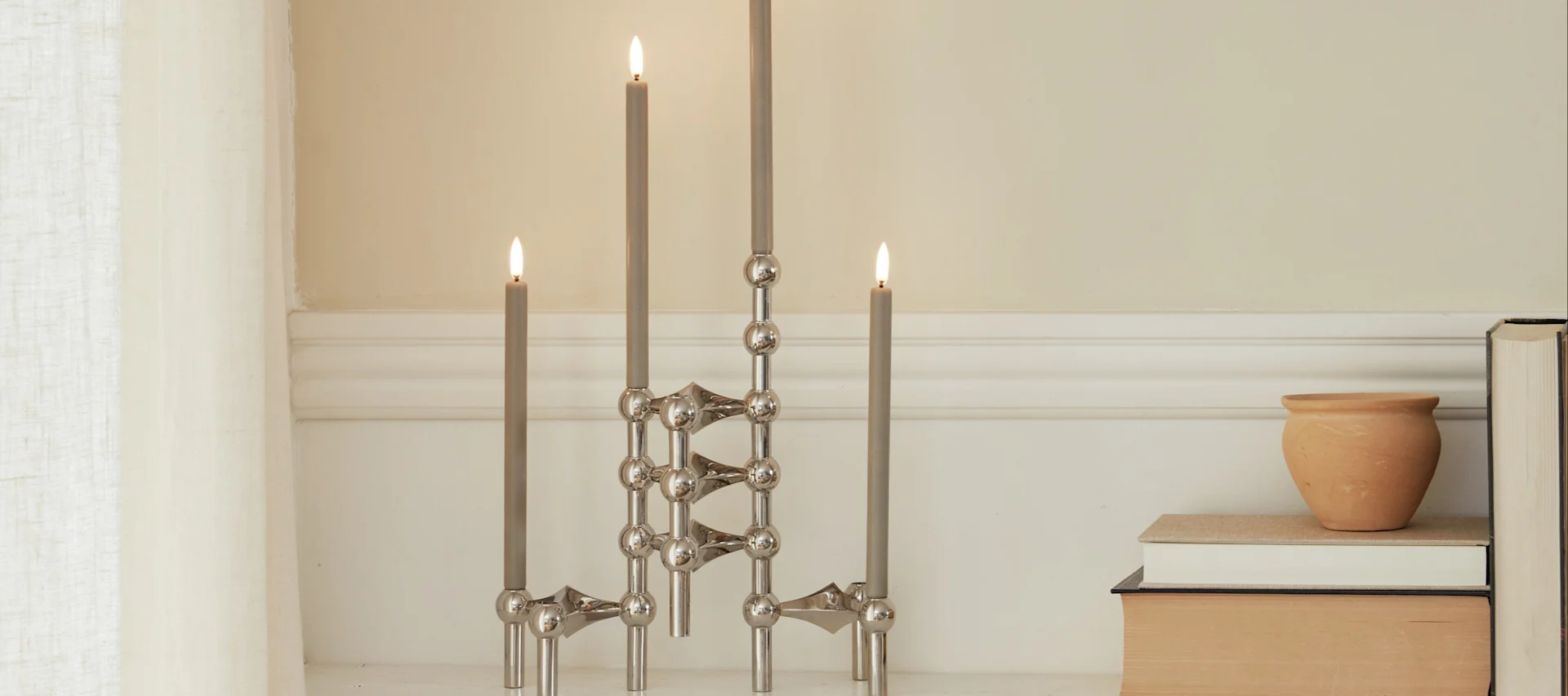 Tres candelabros modulares plateados con velas grises encendidas, junto a una maceta de terracota y libros.