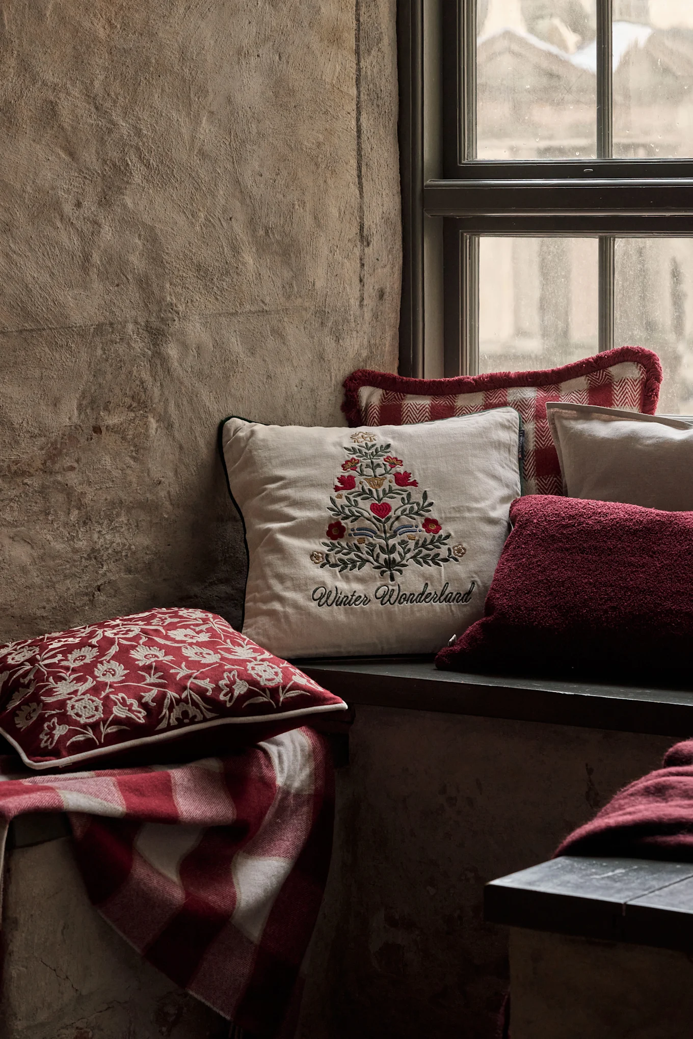 Acogedor rincón de ventana con cojines festivos, incluyendo uno con un árbol de Navidad bordado y 'Winter Wonderland', y una manta a cuadros roja.